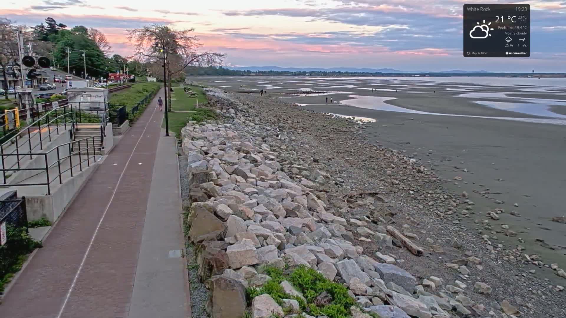 A paved pathway next to a rocky shoreline leads to a mostly cloudy sunset over a wide, exposed tidal flat.