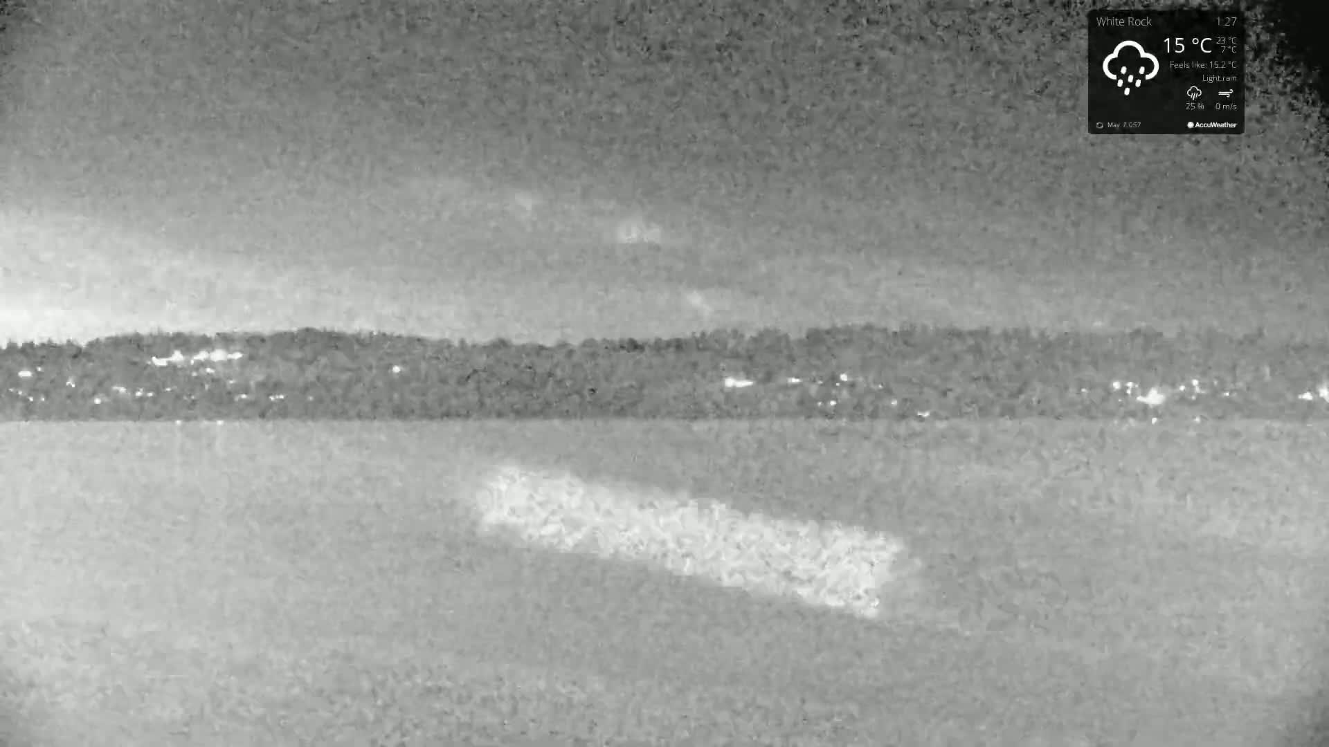 A nighttime, grayscale image shows a body of water with a distant, dimly lit shoreline under a light rain.
