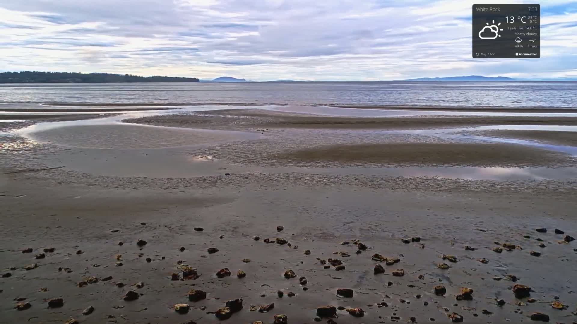 A mostly cloudy day reveals a tidal flat with scattered rocks and pools of water at low tide, overlooking a calm body of water and distant land.