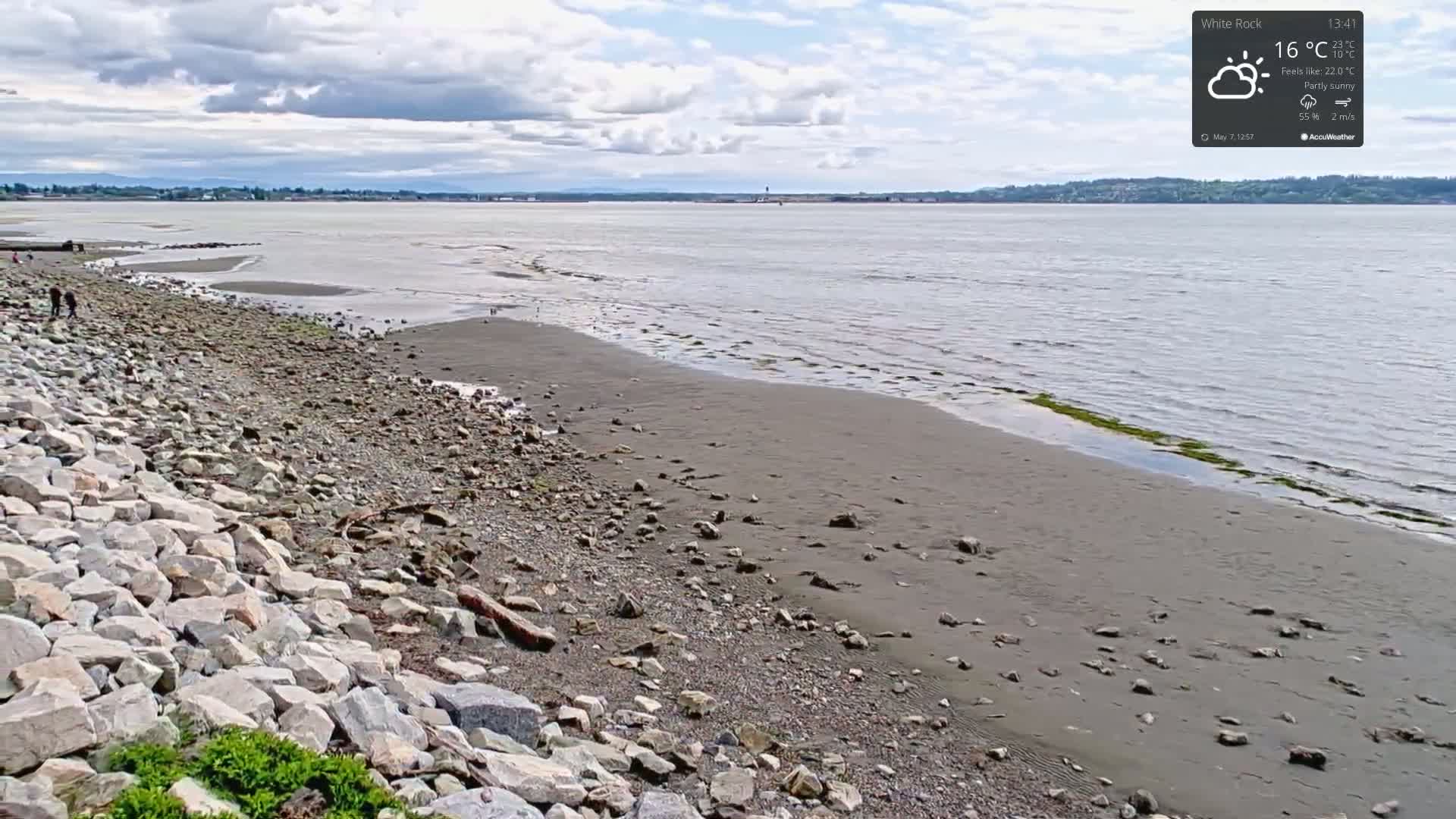A partly sunny day reveals a rocky beach sloping gently toward a calm body of water under a cloudy sky.