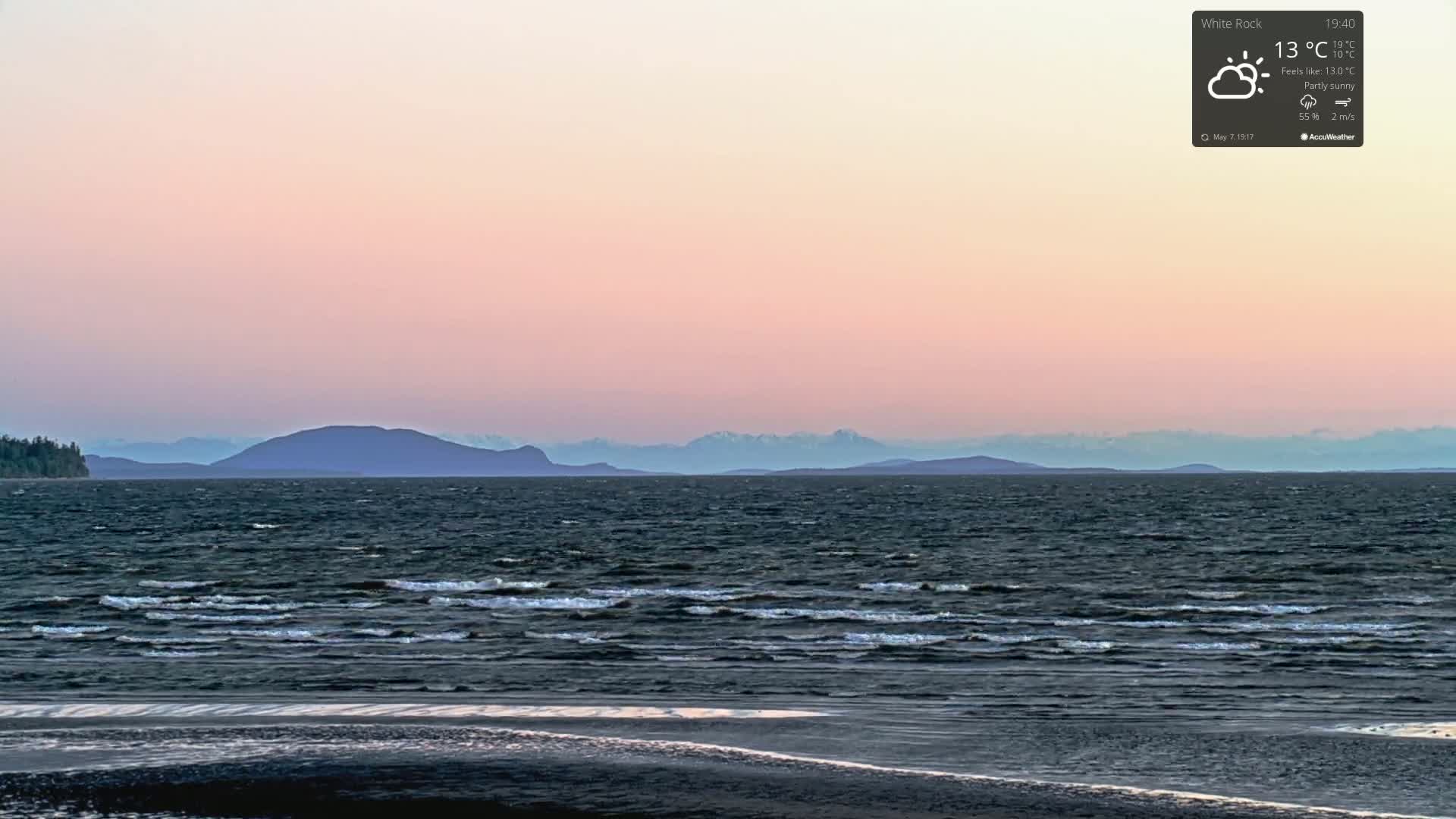A partly sunny, cool evening scene shows a calm body of water with small waves lapping a sandy shore, backed by distant mountains under a pale pink sky.