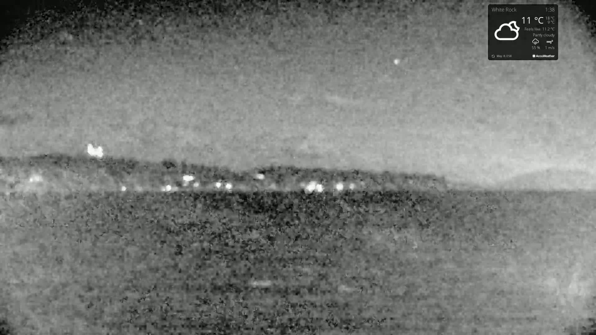 A nighttime, partly cloudy scene shows a dark body of water in the foreground and a distant, dimly lit shoreline.