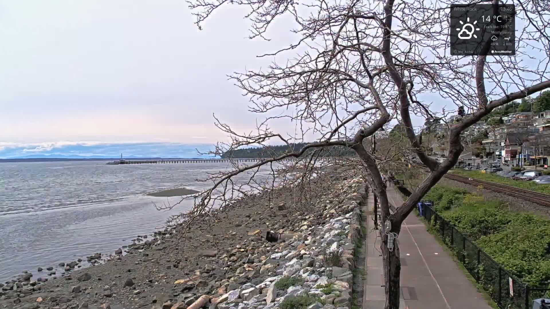 A mostly sunny day shows a rocky shoreline with a walking path next to train tracks and a distant pier, under a mostly bare tree.