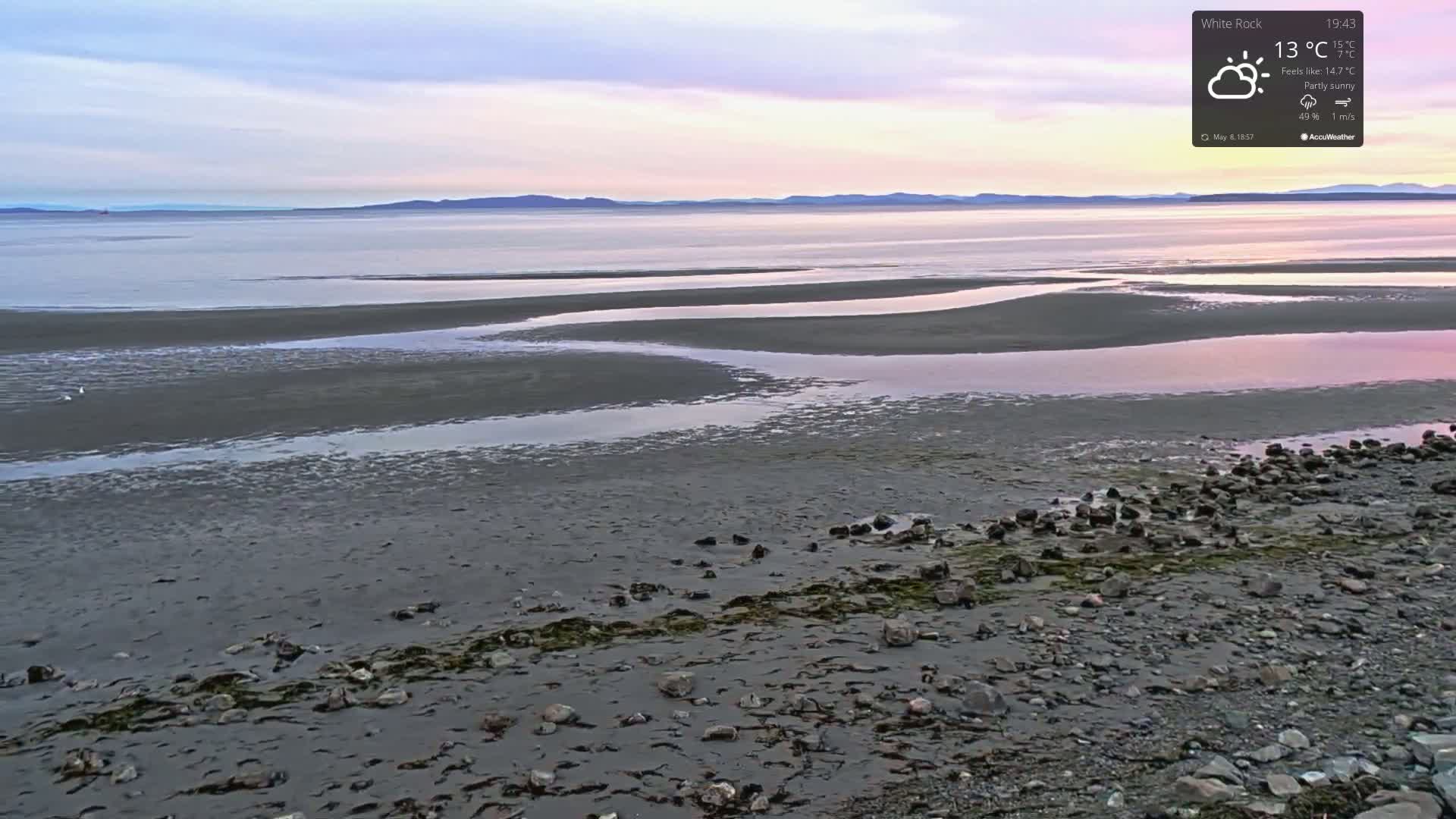 A partly sunny shoreline at low tide reveals a rocky beach and tidal pools under a pale pink and purple sunset sky.