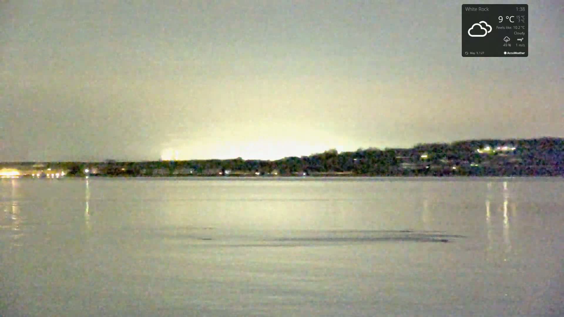 A calm body of water reflects the faint lights of a distant, tree-lined shoreline under a cloudy night sky.