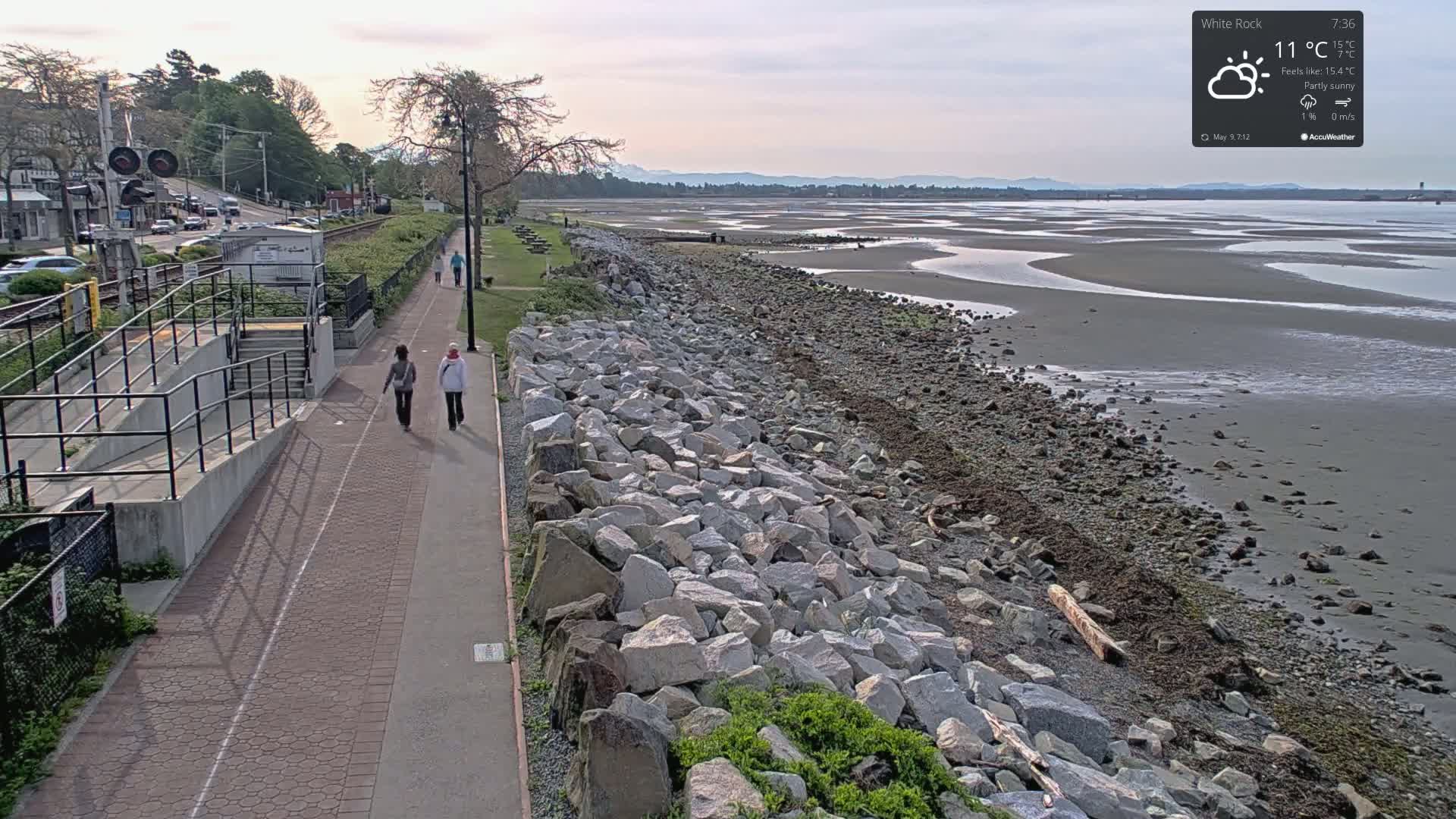 Two people walk along a paved path beside a rocky shoreline, where the low tide reveals a wide expanse of wet sand under a partly sunny sky.