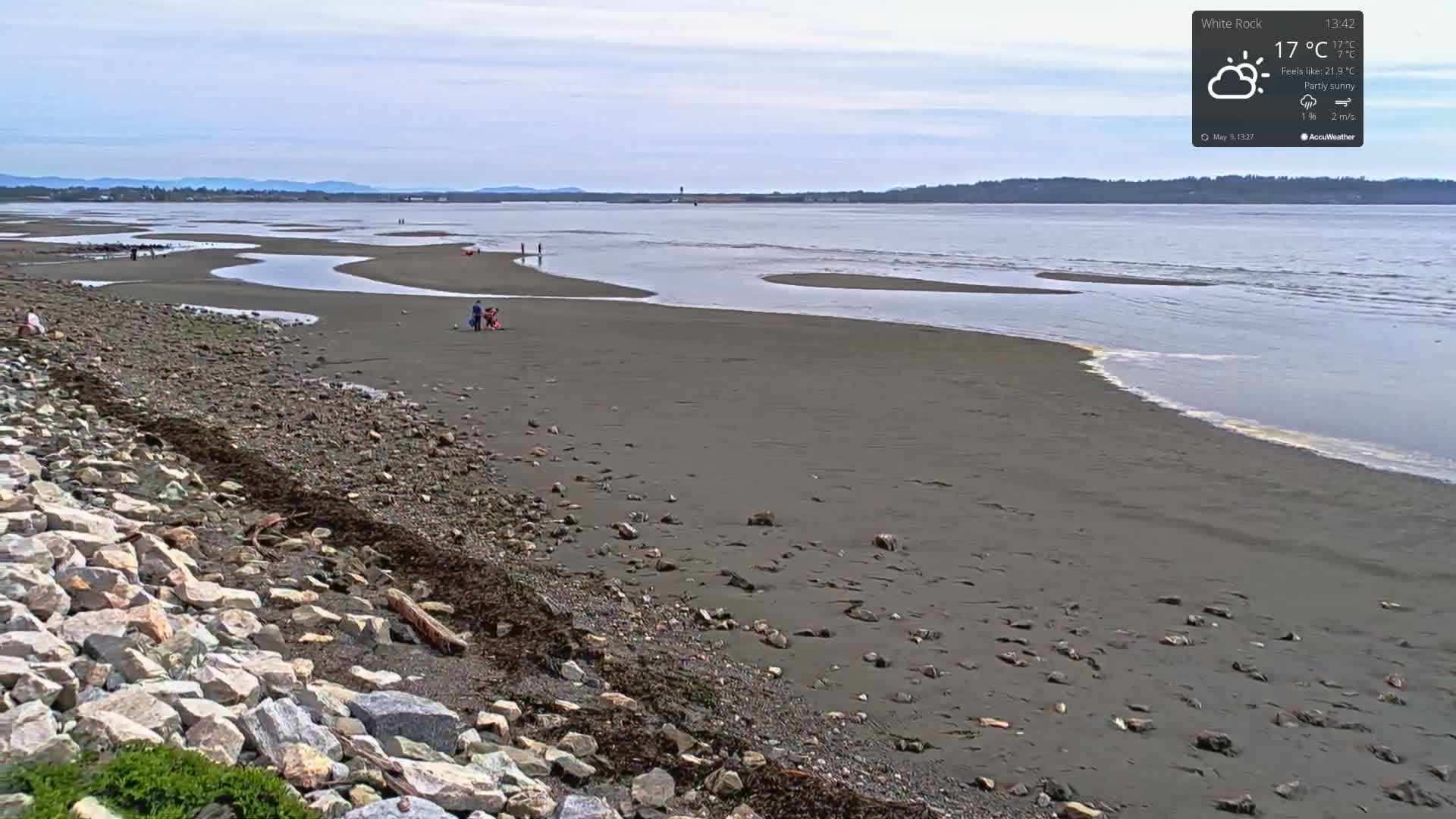 A partly sunny day at a rocky beach with a calm body of water and several people scattered on the shore.