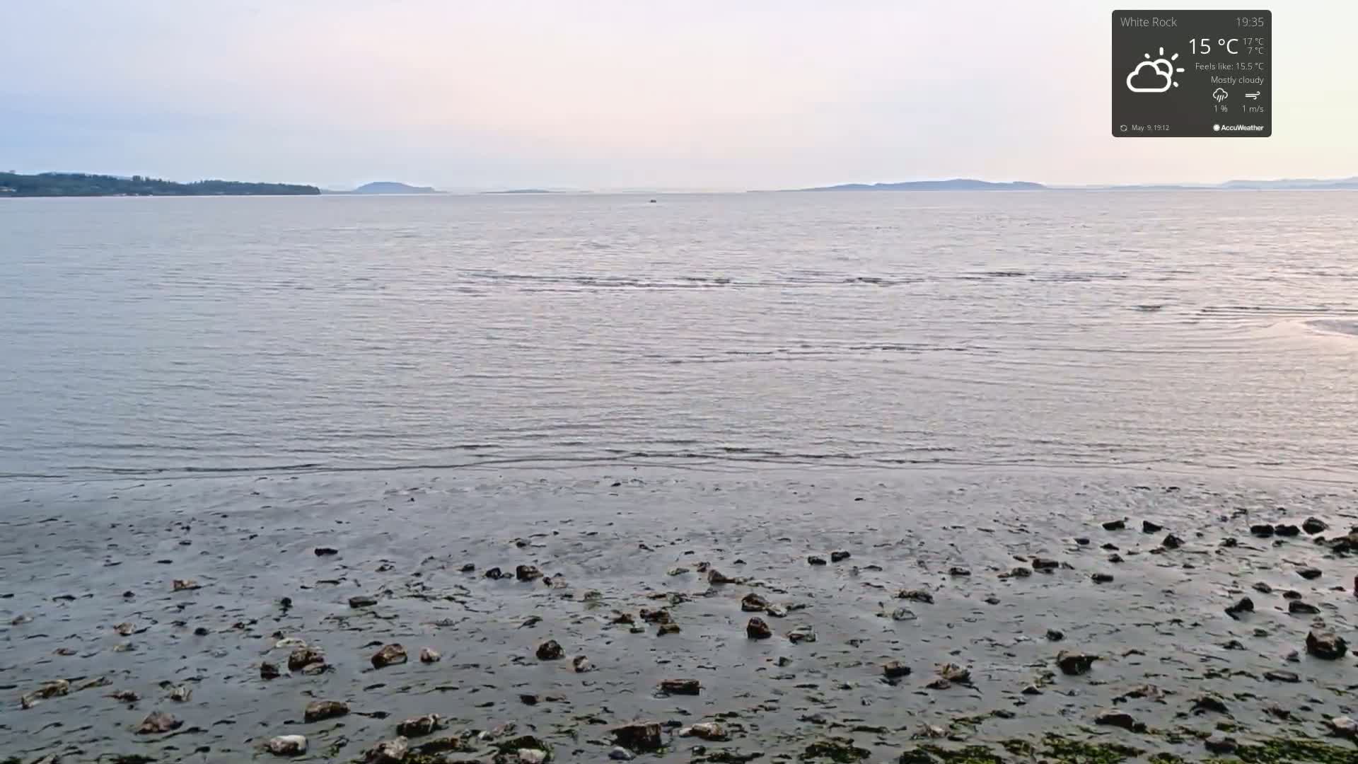 A mostly cloudy, calm day reveals a shoreline with scattered rocks at low tide, overlooking a wide expanse of calm water and distant land.
