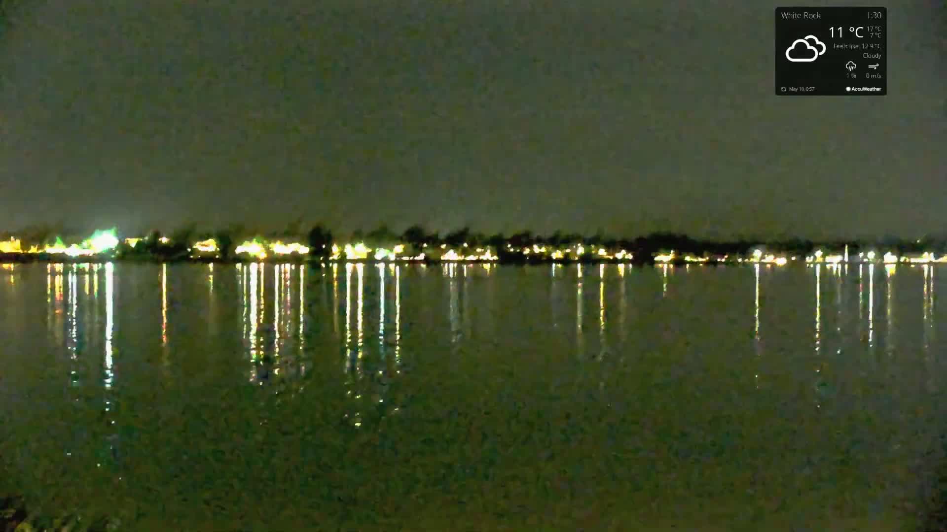 At night, under cloudy skies, the lights of a town are reflected in a calm body of water.