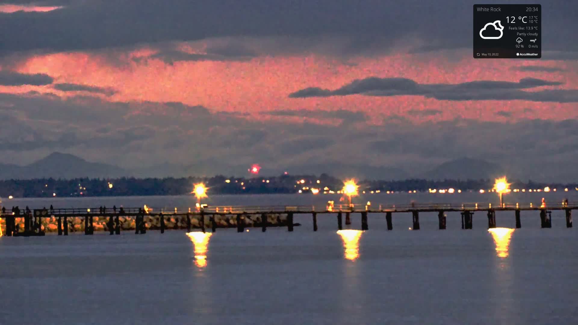 A long pier with people on it is illuminated at night under a partly cloudy sky with pink and purple hues in the twilight.