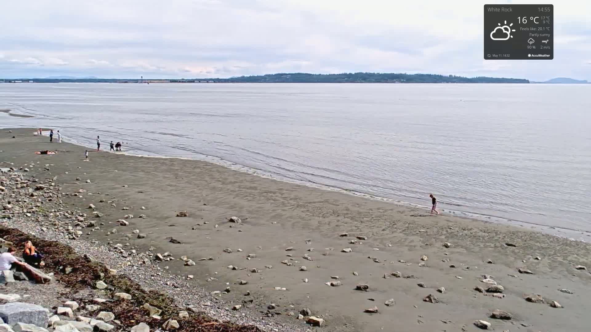 A mostly sunny day shows a wide sandy beach with scattered rocks, a calm body of water, and several people enjoying the shore.
