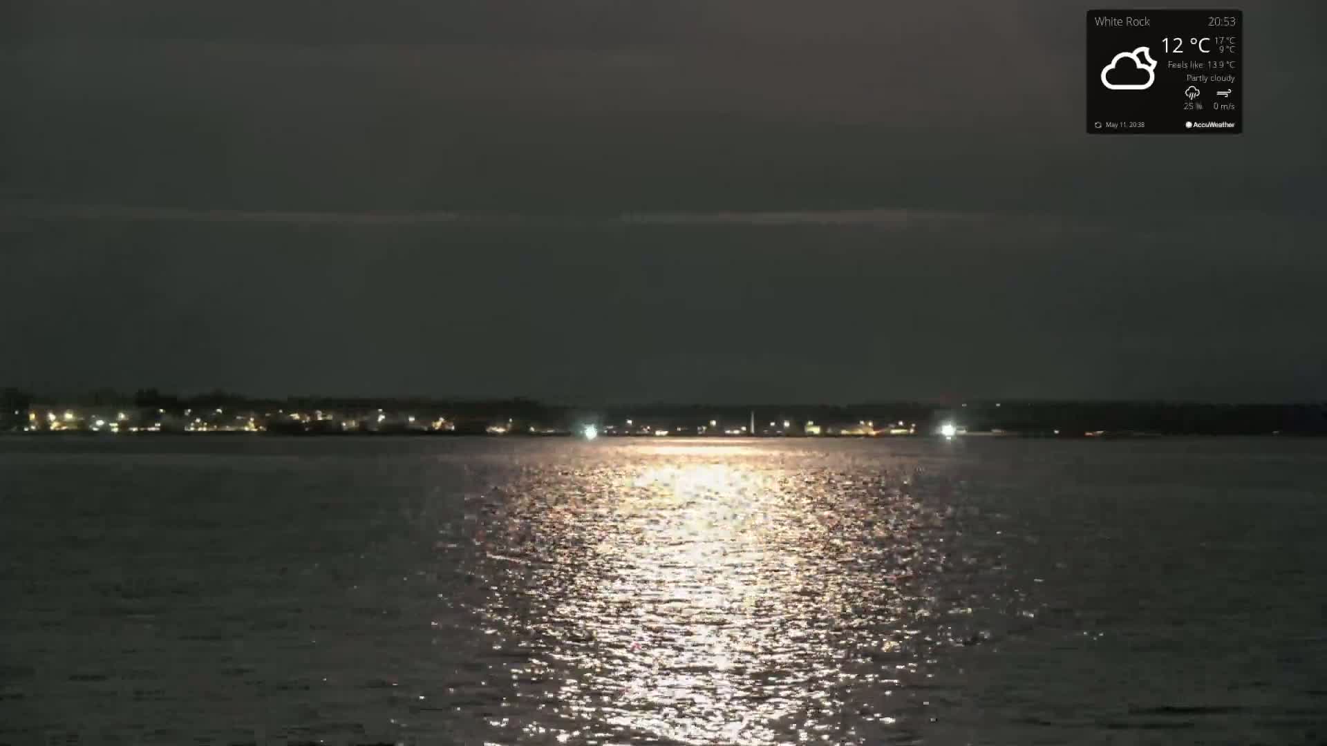 A moonlit, partly cloudy night scene shows calm water reflecting the lights of a distant shoreline.