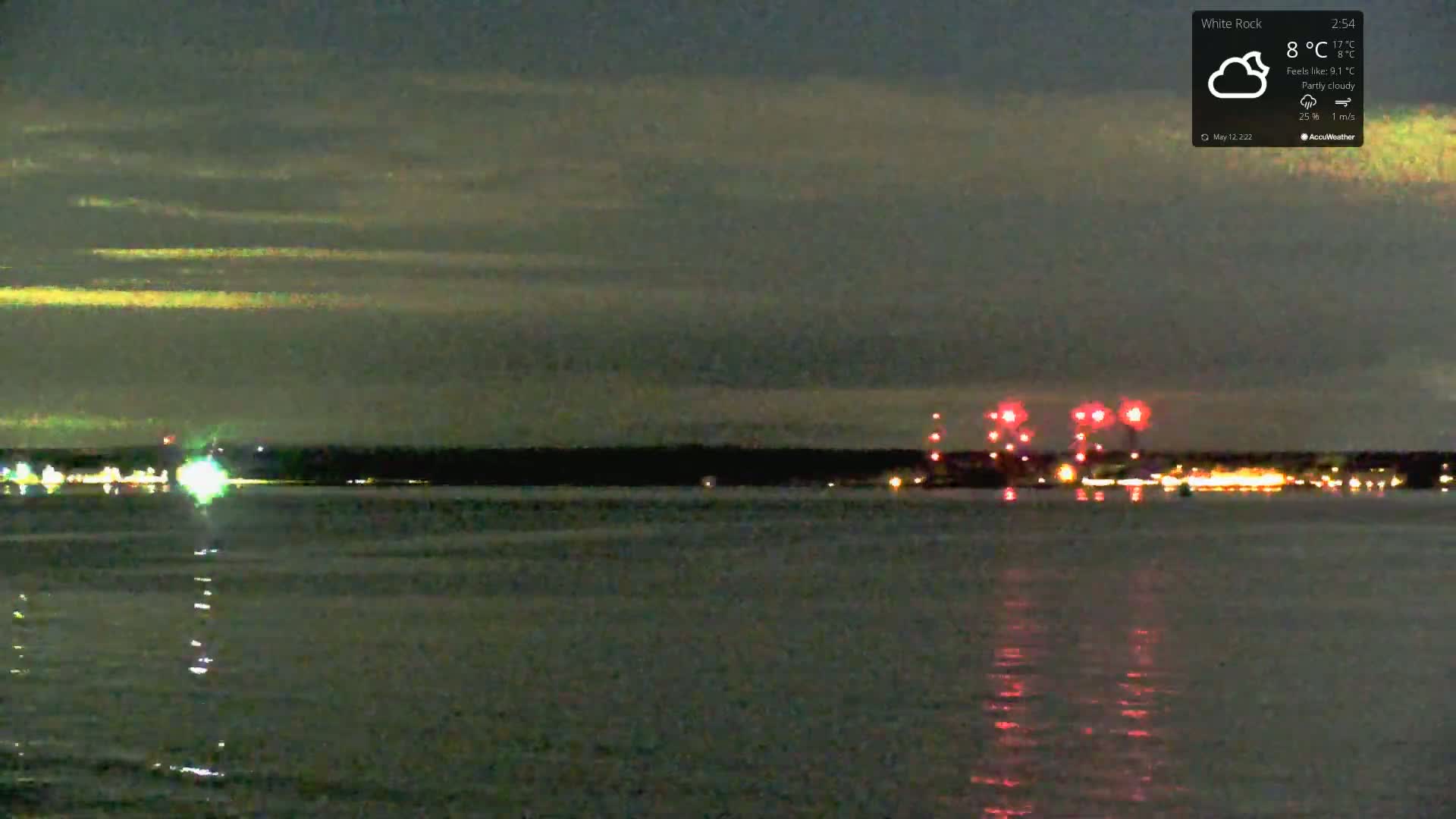 A nighttime view of a calm body of water with fireworks exploding over a distant shoreline under a partly cloudy sky.