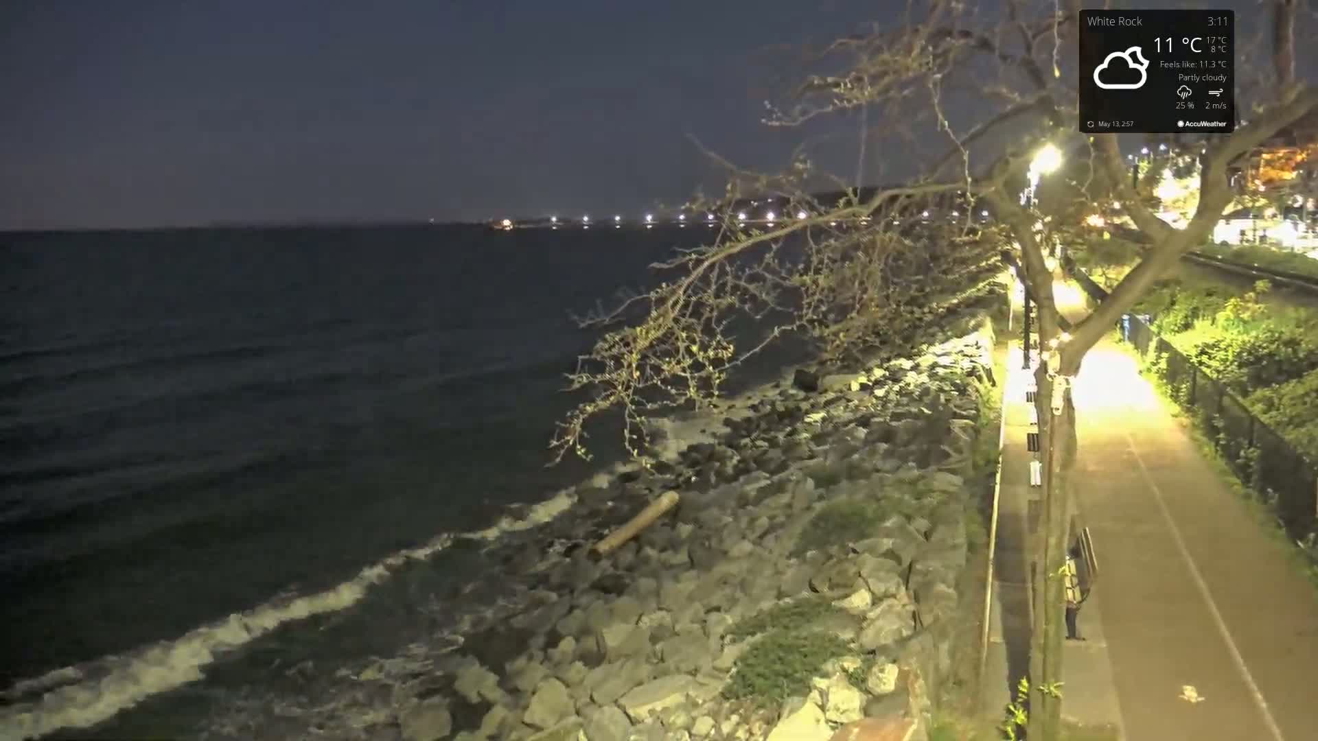 A partly cloudy night scene shows a rocky shoreline with gentle waves lapping against it, a paved path lit by streetlights, and a person walking along the path.