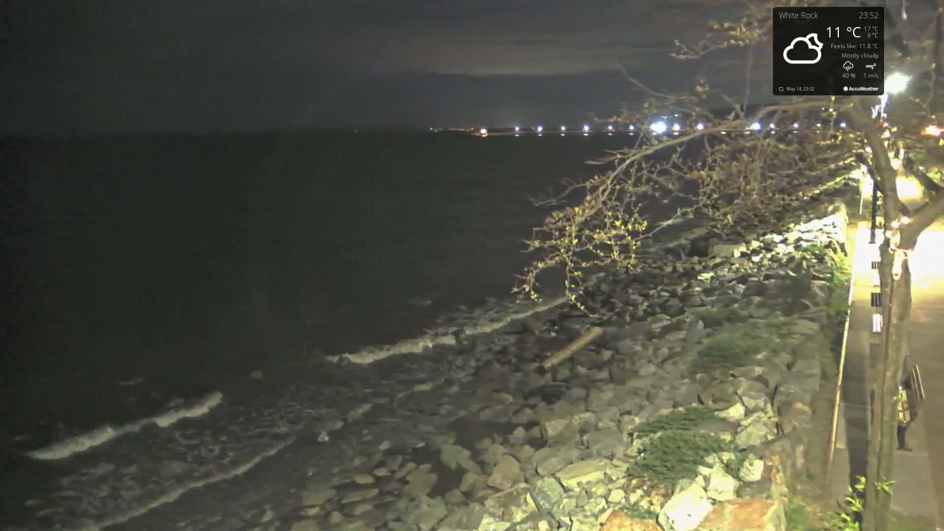 A dark, mostly cloudy night scene shows a rocky shoreline with small waves lapping at the rocks, and lights visible in the distance across the water.