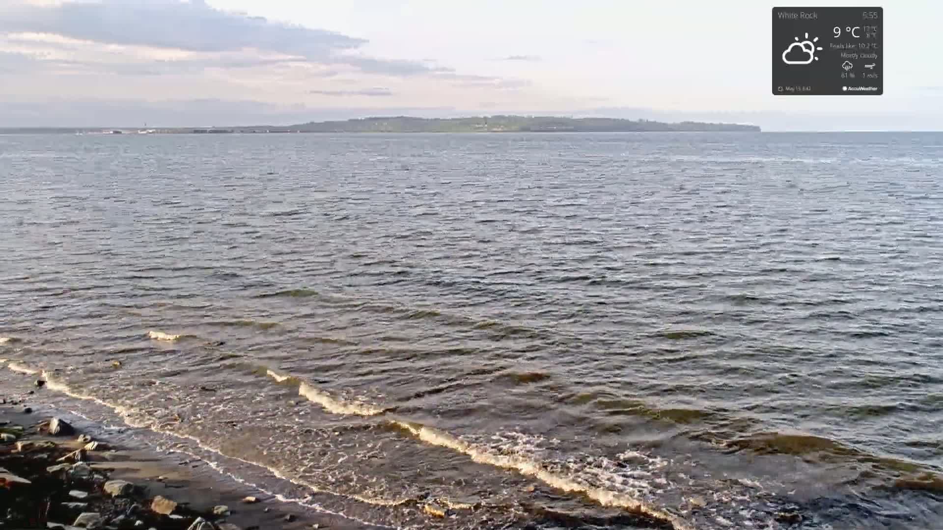 Small waves lap a rocky shoreline on a mostly cloudy day, with a distant landmass visible across a body of water.