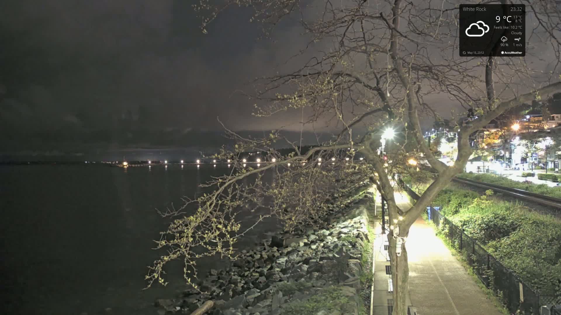 A nighttime view of a rocky shoreline with a tree and a paved path next to train tracks under cloudy skies.