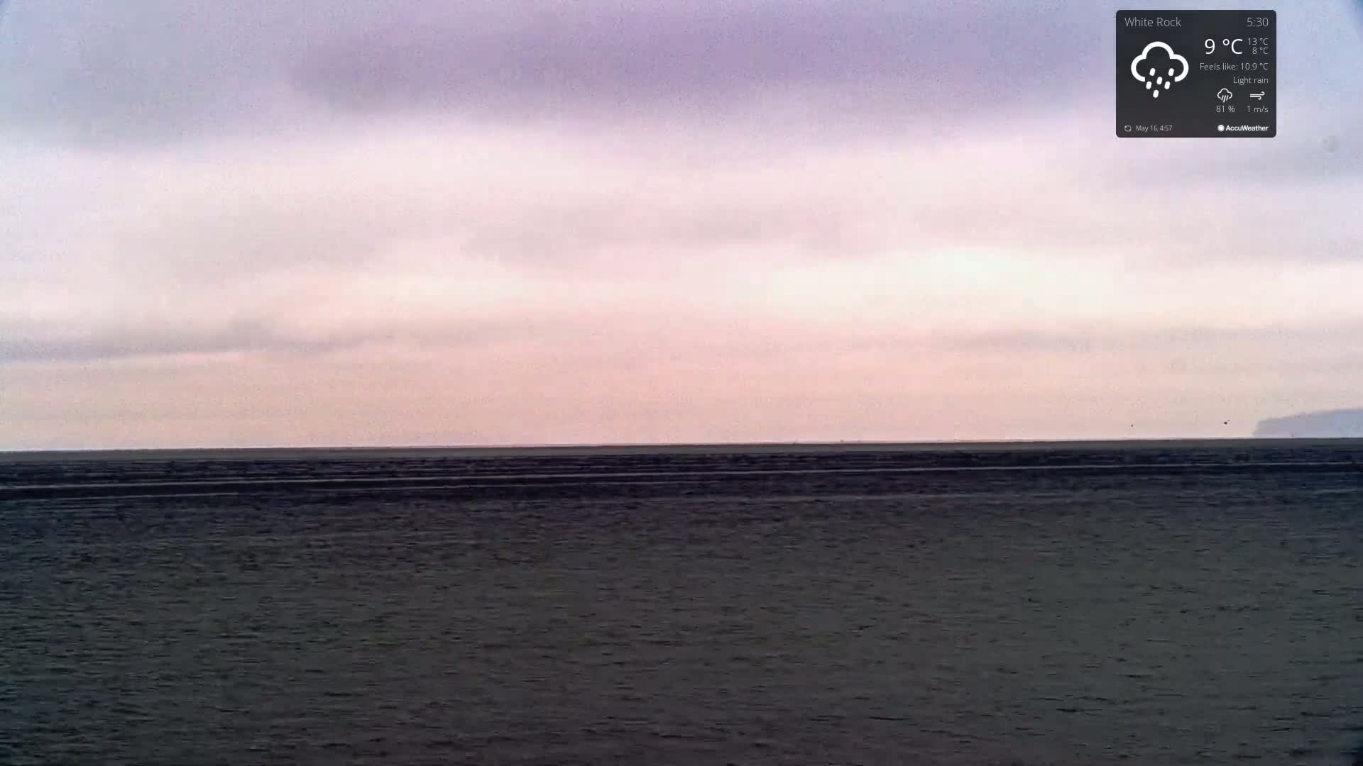A dark, flat body of water extends to a pale pink and gray horizon under a lightly overcast sky with light rain.