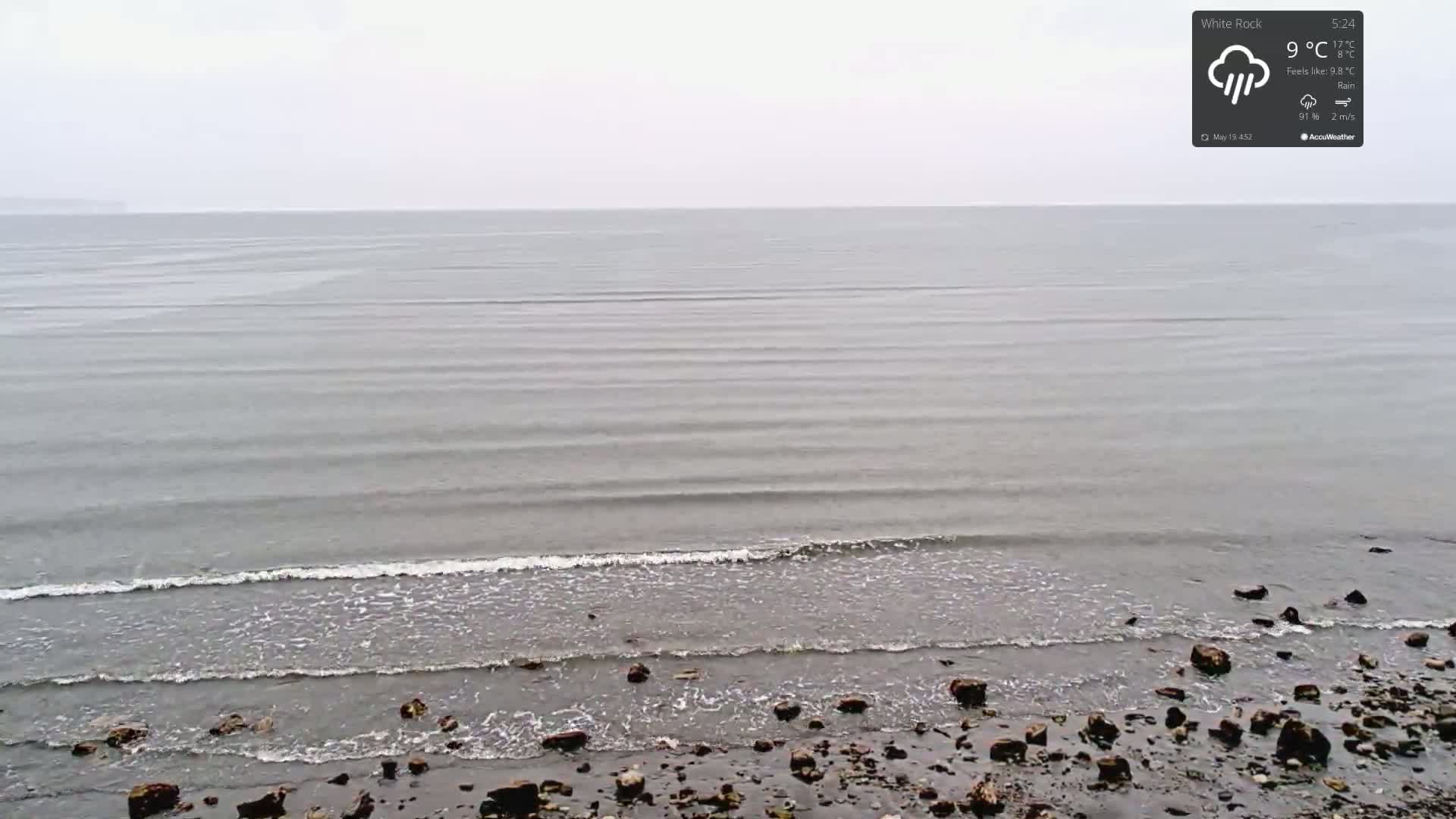A rocky shoreline meets a calm, gray sea under a rainy sky.