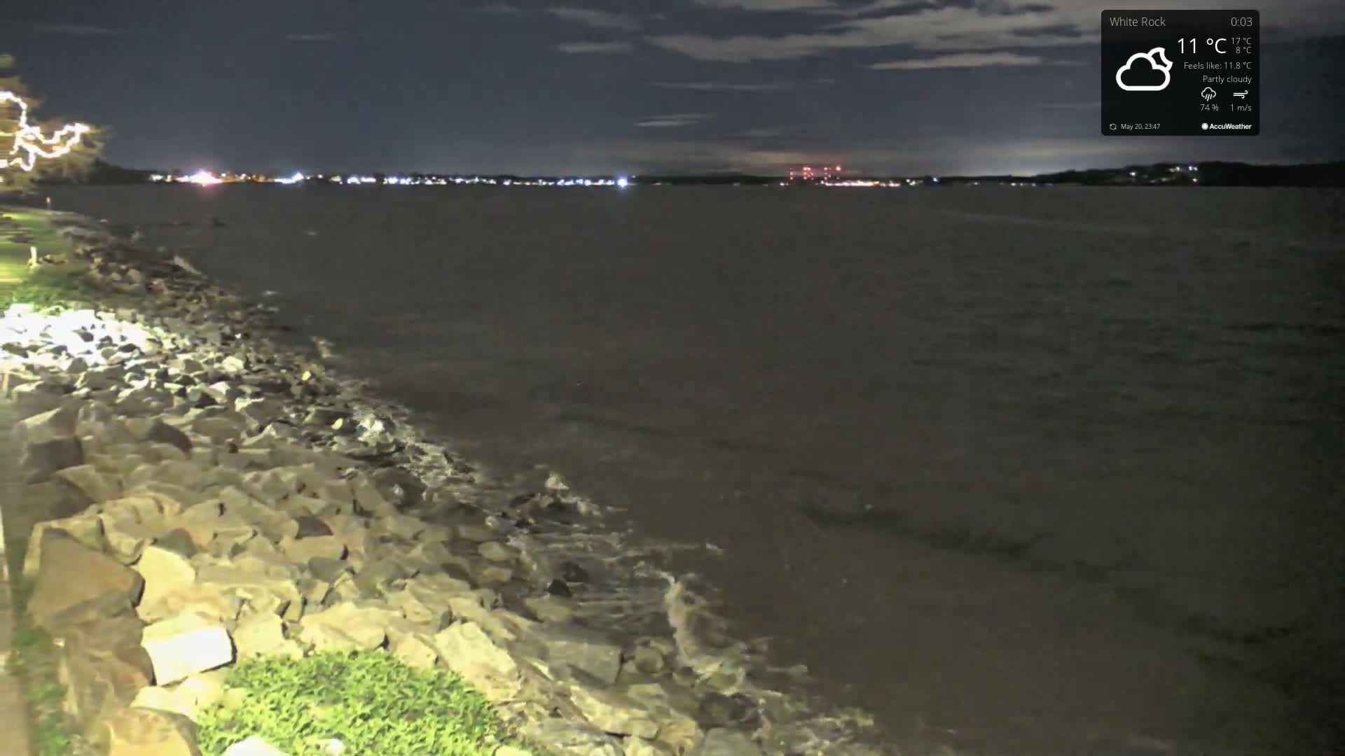 A rocky shoreline at night under a partly cloudy sky meets a dark body of water with small waves, and distant city lights are visible on the opposite shore.