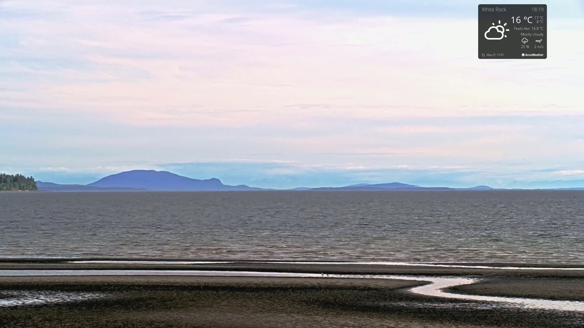 A mostly cloudy day with a view of a calm bay and distant mountains from a sandy beach.