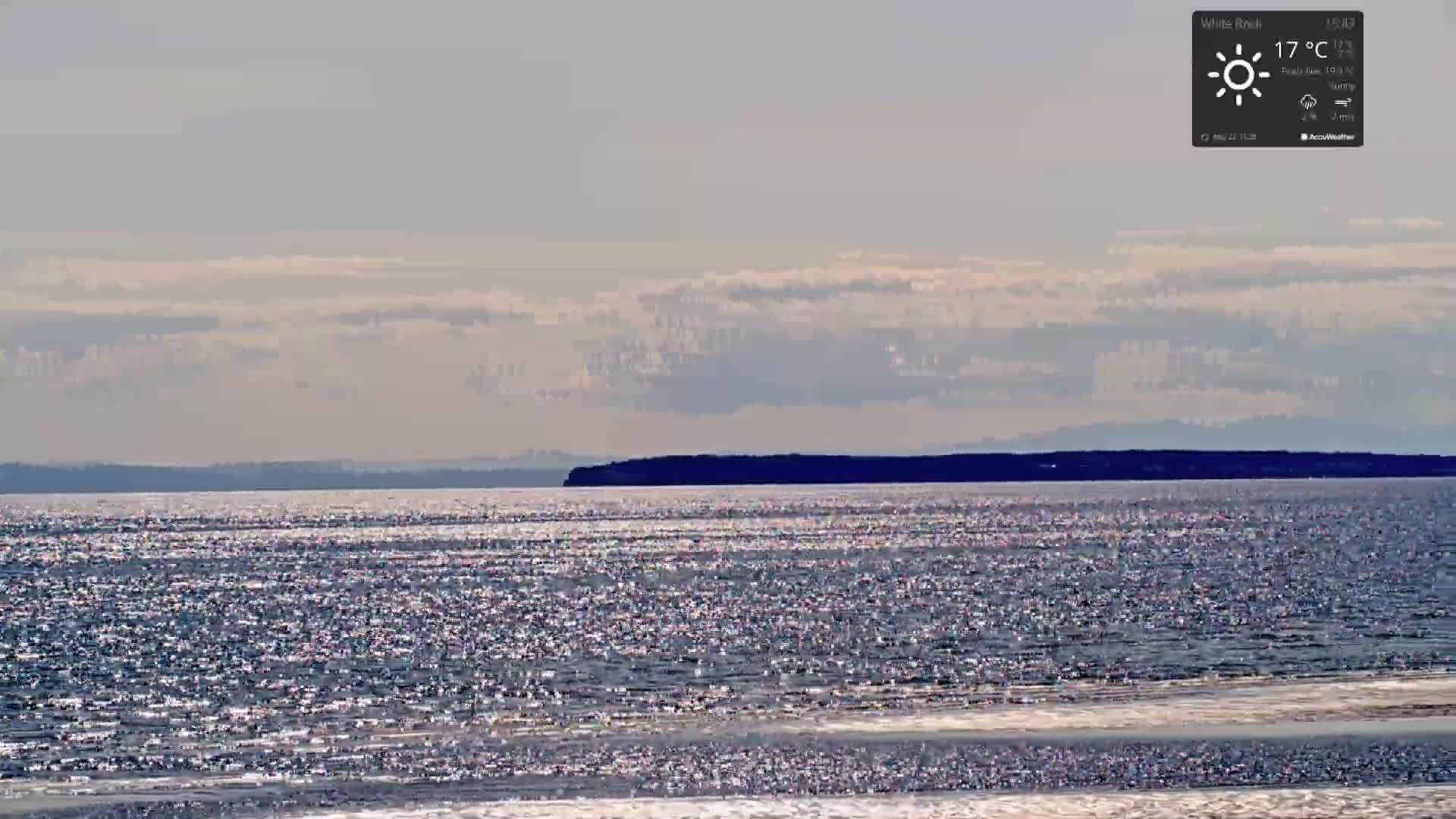A mostly sunny day shows a calm body of water sparkling under a mostly cloudy sky, with a dark landmass visible in the distance.