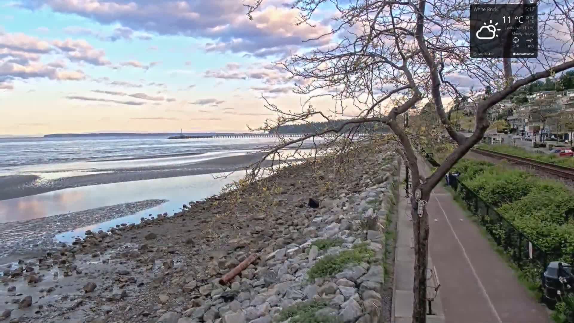 A mostly cloudy sky overlooks a rocky shoreline with a calm body of water, a pathway, and a railway track beside a residential area.