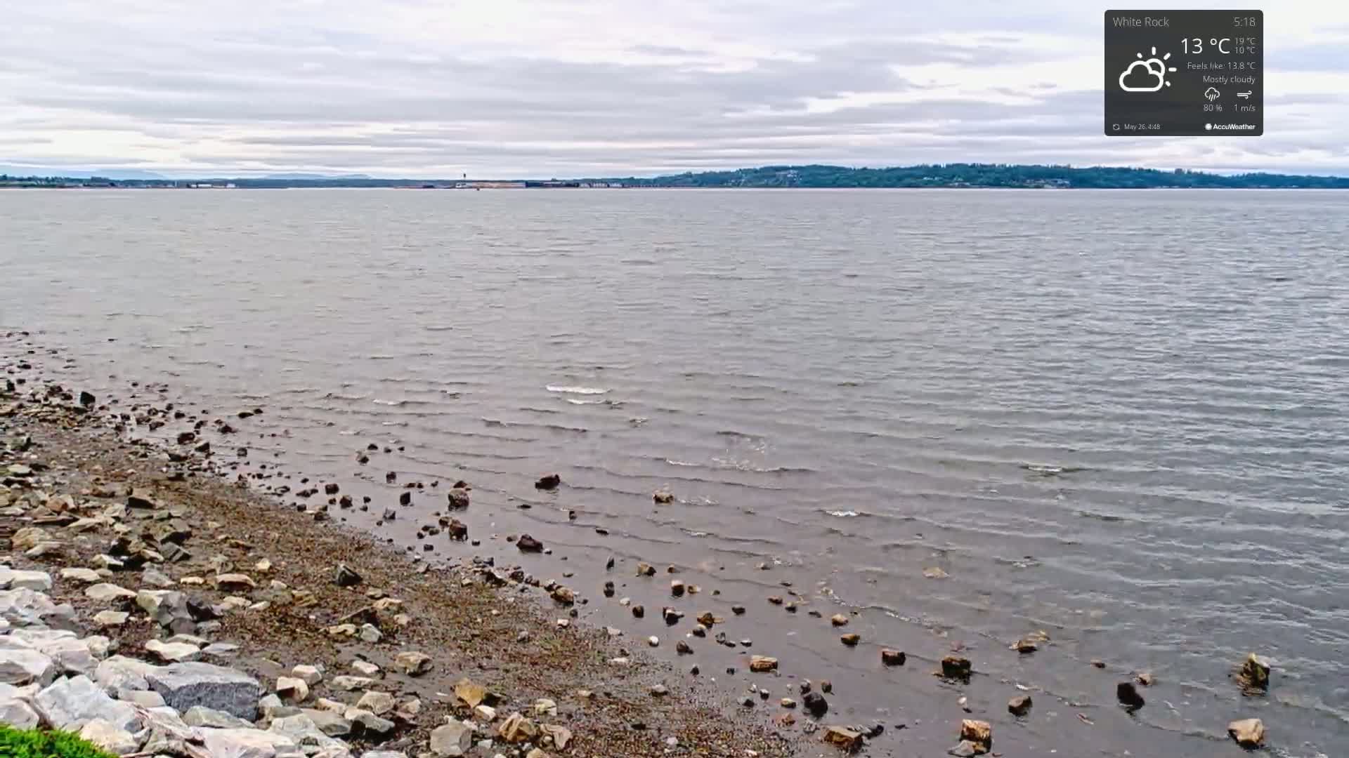 A rocky shoreline meets a calm, gray body of water under a mostly cloudy sky.