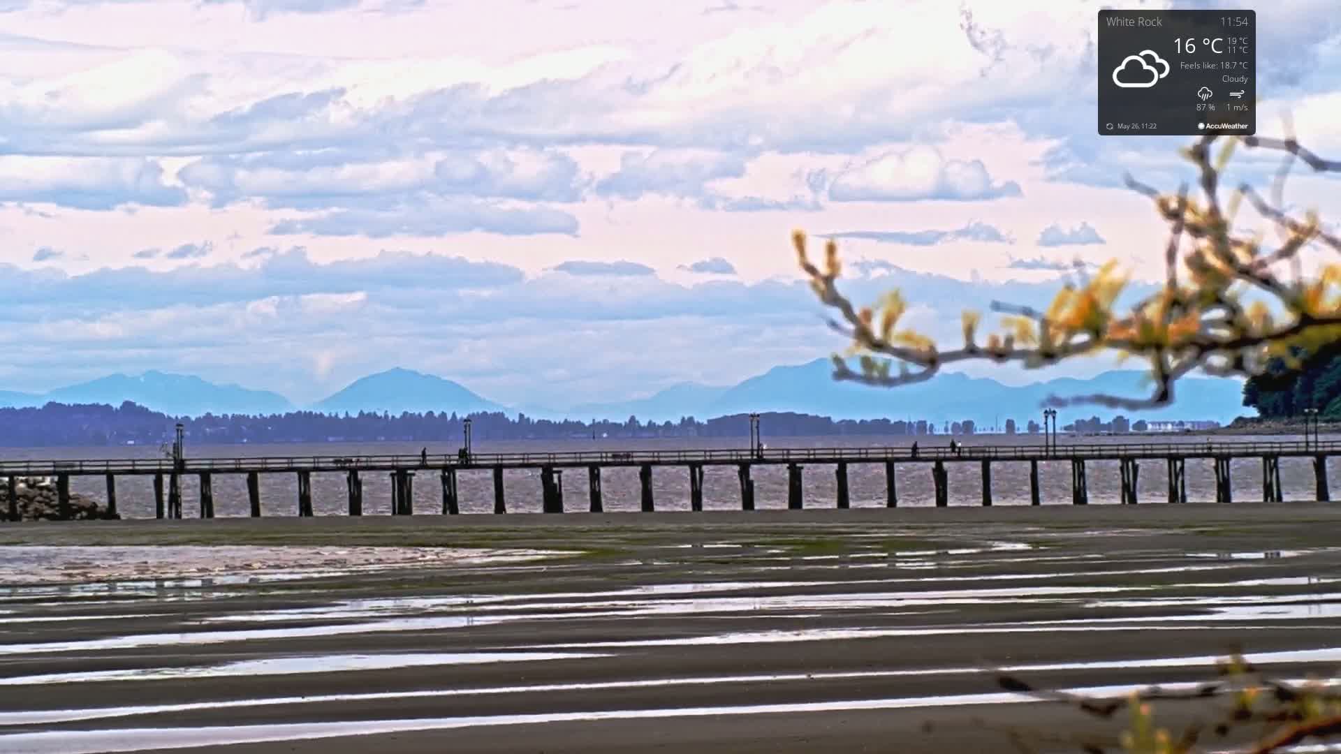A long pier stretches across calm water towards a distant mountain range under a partly cloudy sky.