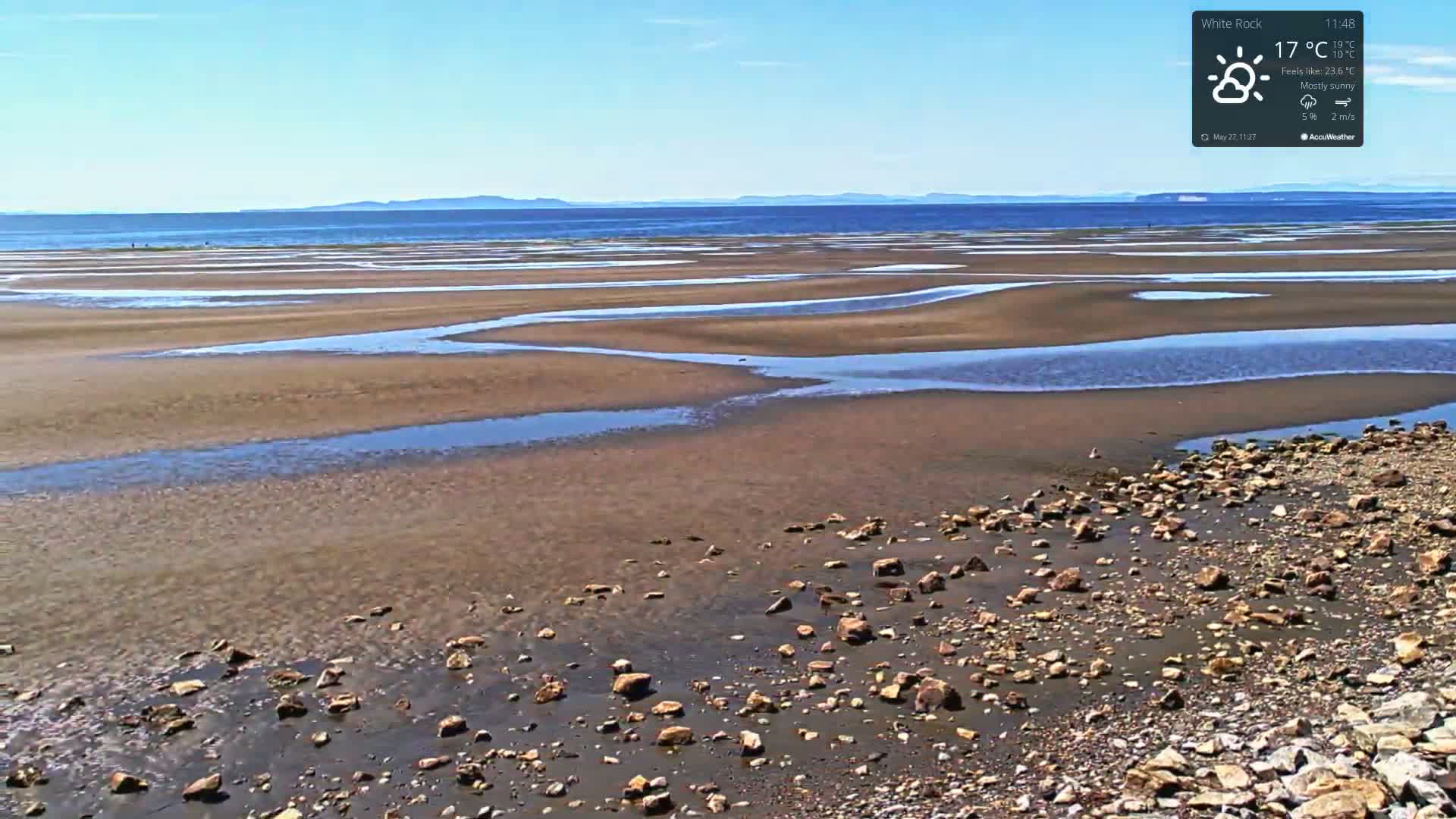 A mostly sunny day reveals a wide expanse of sandy beach with shallow pools of water at low tide, and a rocky shoreline in the foreground, under a clear blue sky and distant mountains.