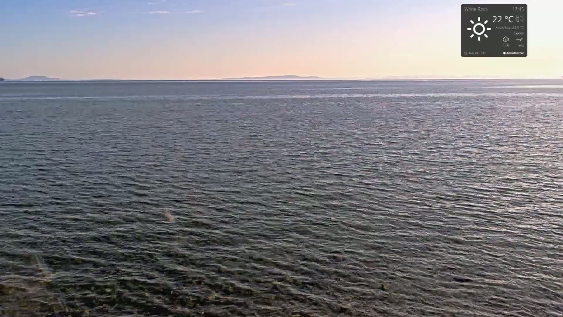 A calm, sunny body of water stretches to a distant, low-lying landmass under a clear, light blue sky.