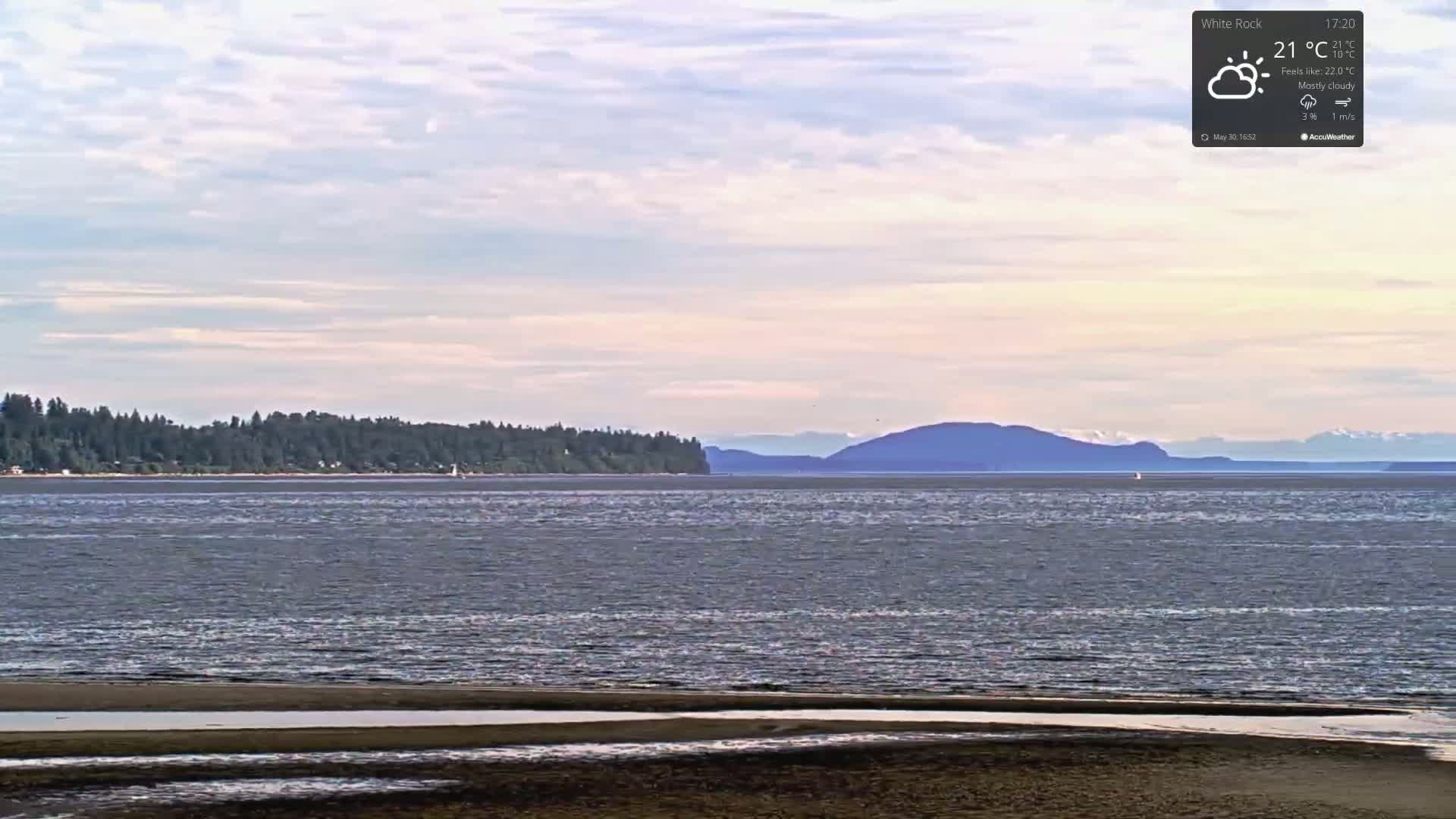 A mostly cloudy day reveals a tranquil body of water with a sandy shoreline in the foreground and a distant treeline and mountains under a pale sky.