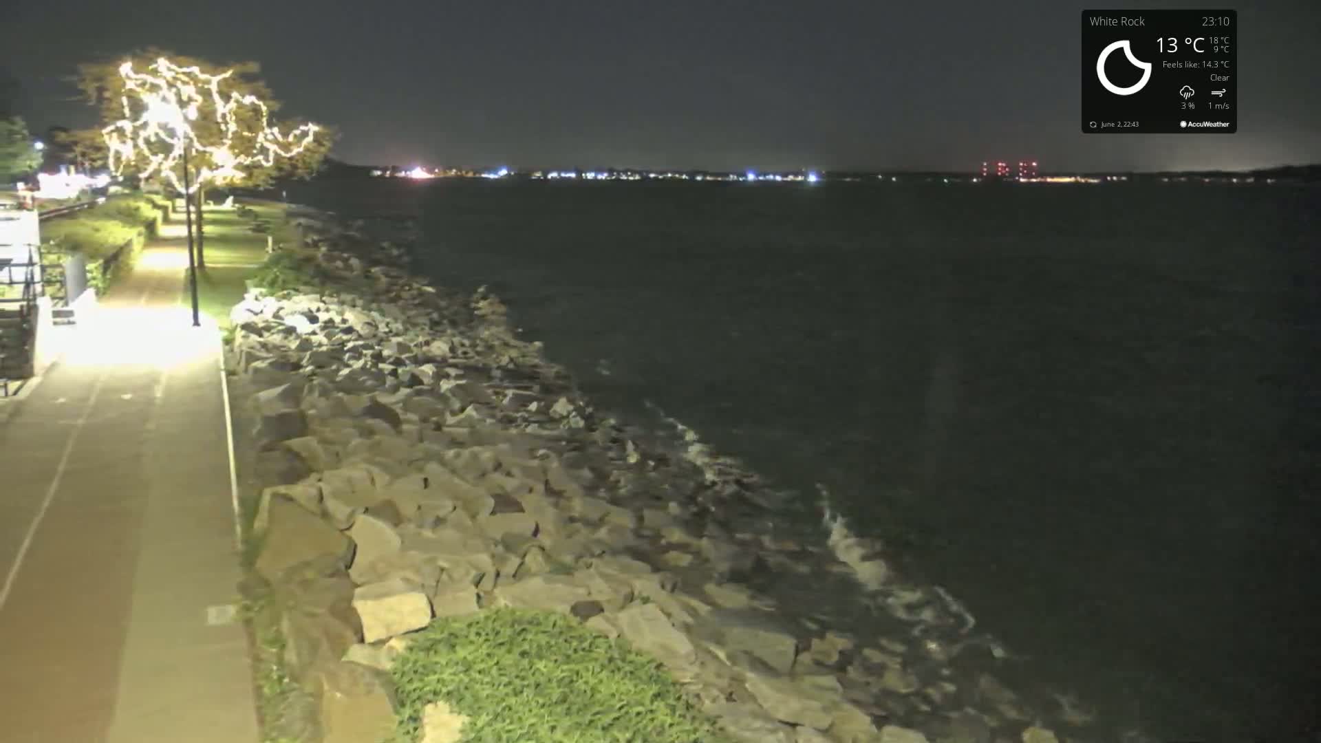 A nighttime view of a paved walkway next to a rocky shoreline with gentle waves and a city skyline faintly visible in the distance under clear skies.
