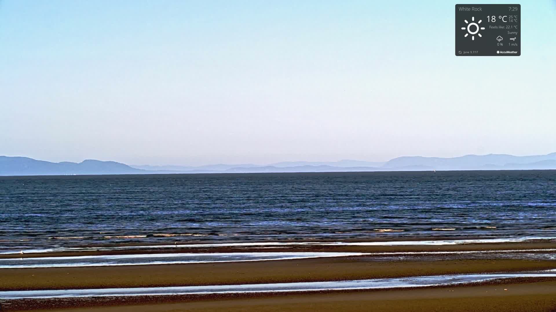 A sandy beach with calm water and distant mountains is visible under sunny skies.