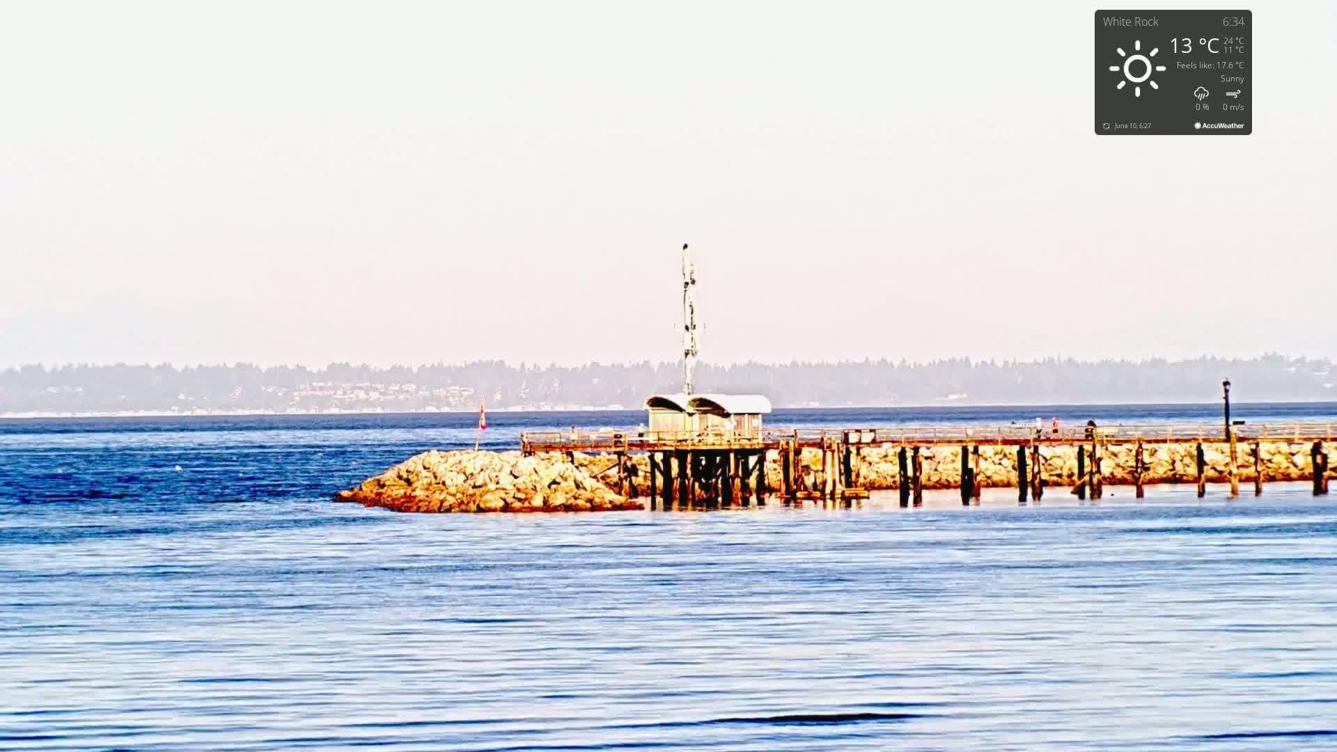 A small structure sits on a pier extending from a rocky outcrop into calm, blue water under a sunny sky, with a hazy tree-lined shore visible in the distance.
