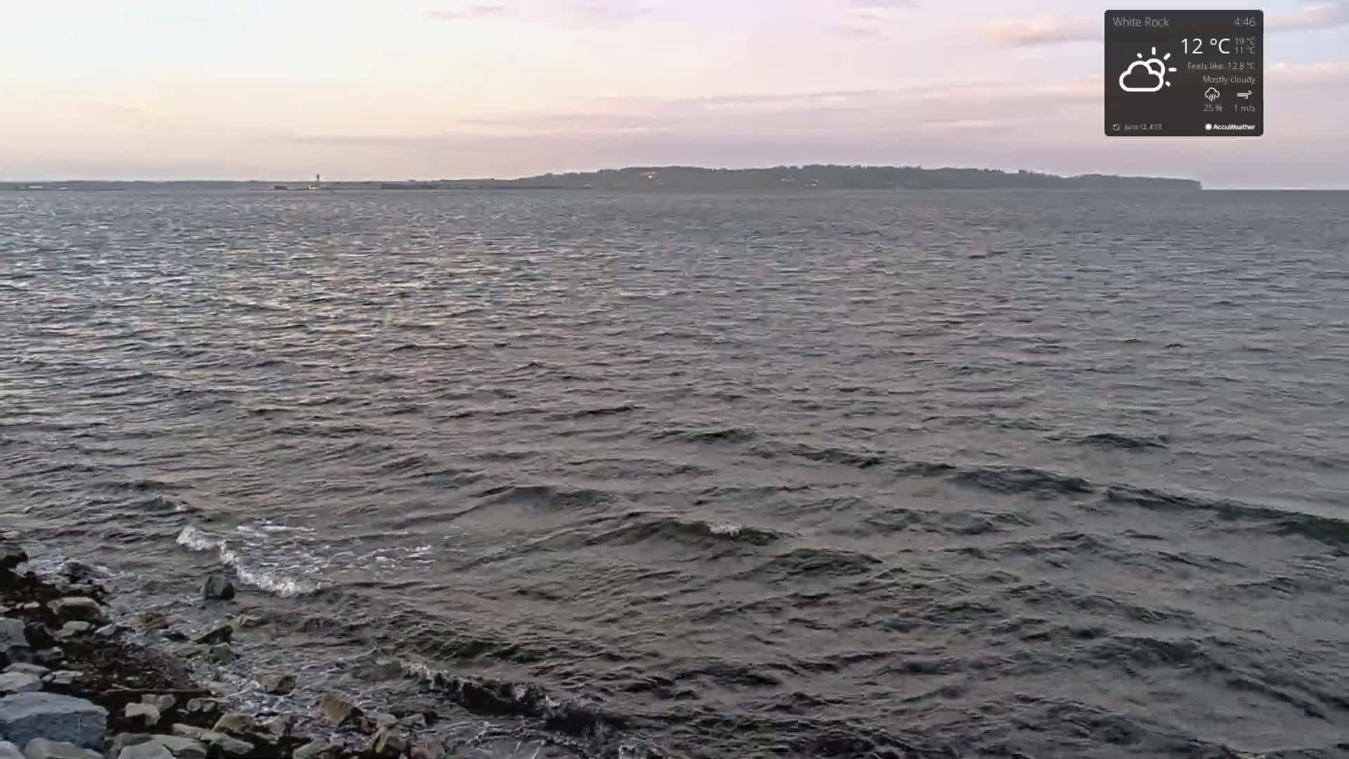 A mostly cloudy sky overlooks a body of water with small waves lapping against a rocky shore.