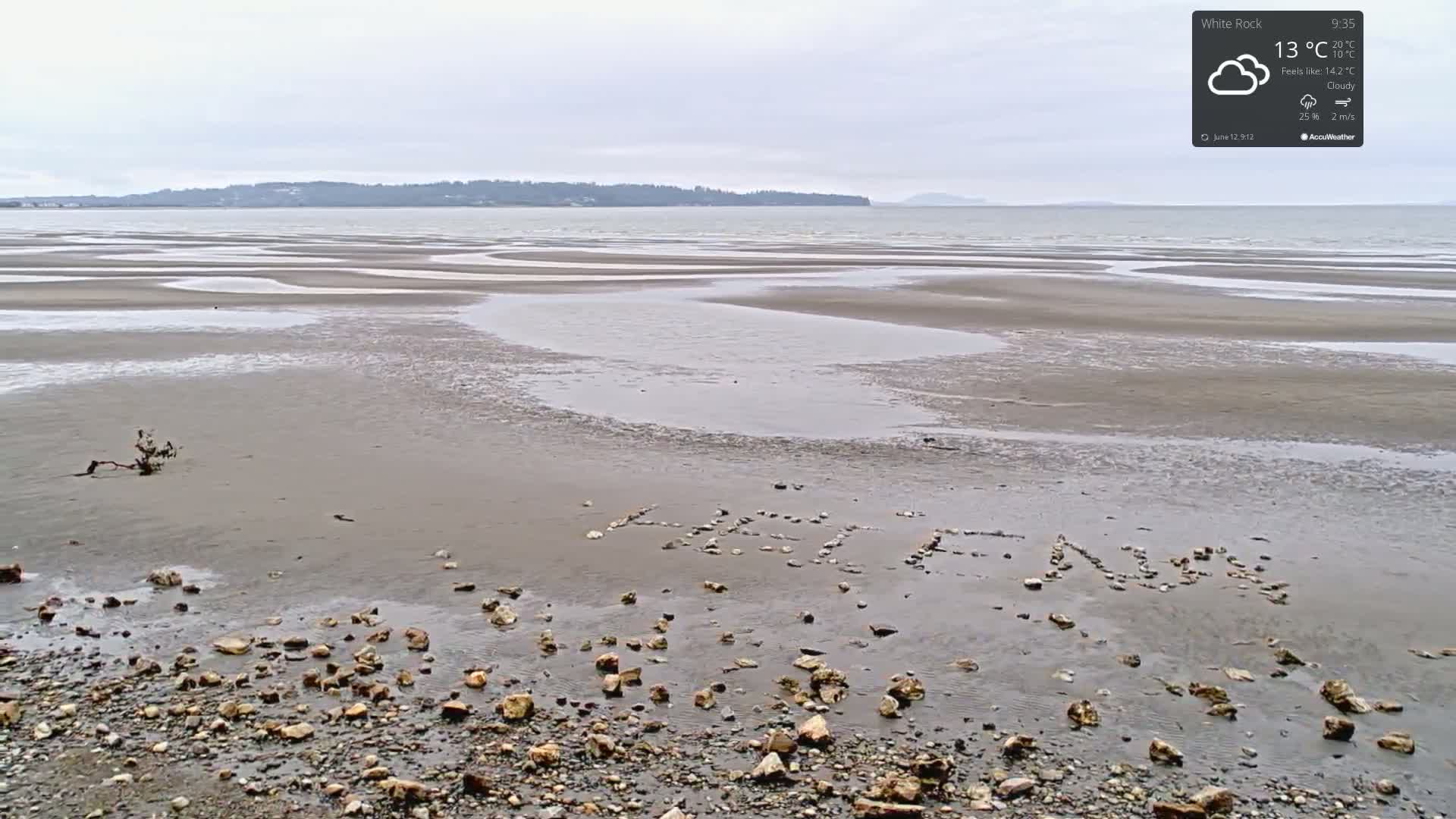 A cloudy day reveals a wide, flat beach with low tide, showing sand patterns and the word "HELEN" spelled out in small stones.