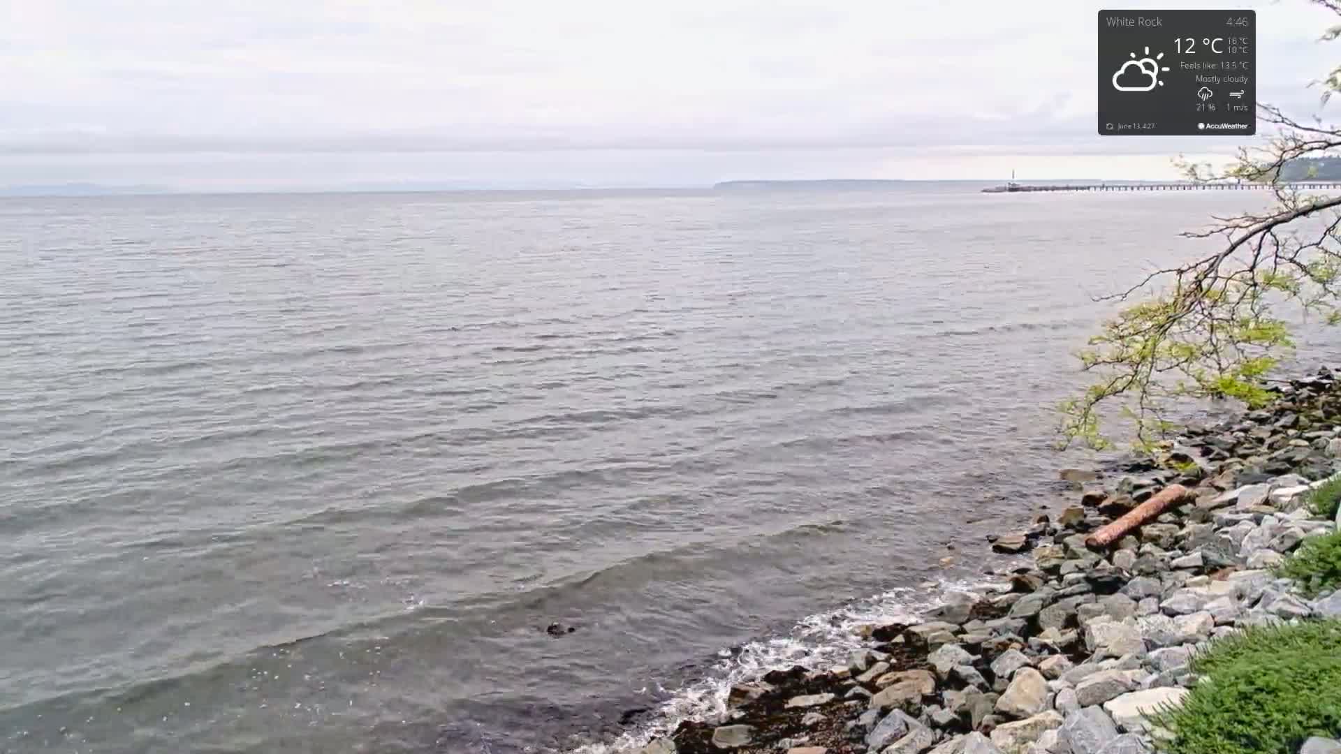 A mostly cloudy day shows a rocky shoreline with gentle waves lapping at the shore, and a calm body of water extending to a distant landmass.