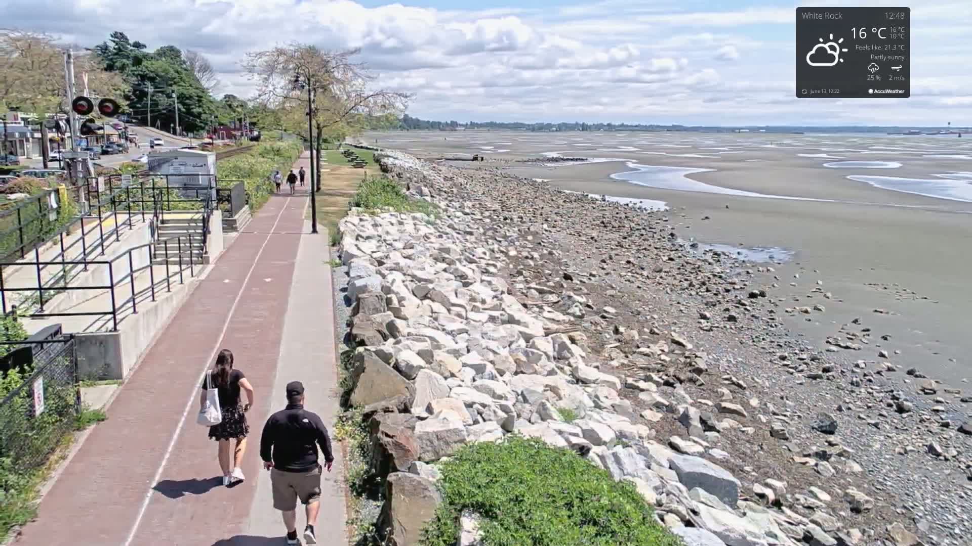 A paved pathway next to a rocky shoreline overlooks a partly sunny beach with exposed mudflats at low tide, and two people walk along the path.