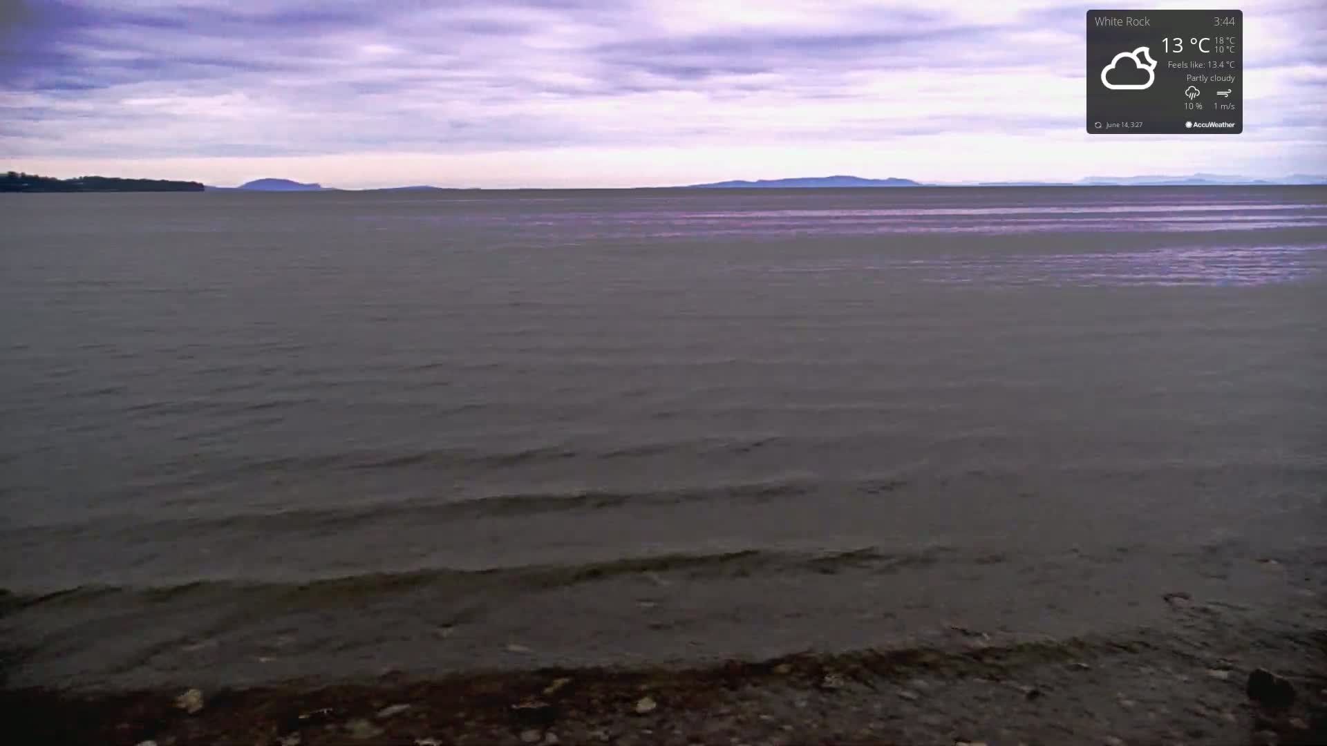 A partly cloudy sky overlooks a calm body of water lapping a dark, pebbly shore.