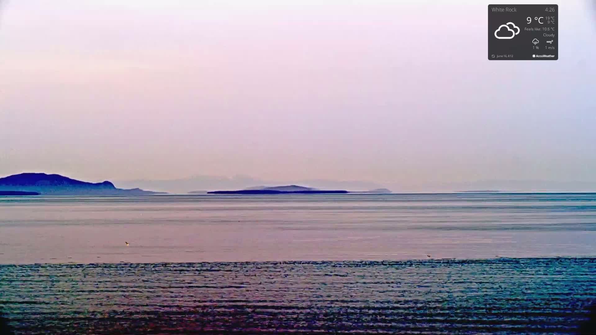 A calm body of water stretches to a distant shoreline of low-lying mountains under a cloudy, pale sky.