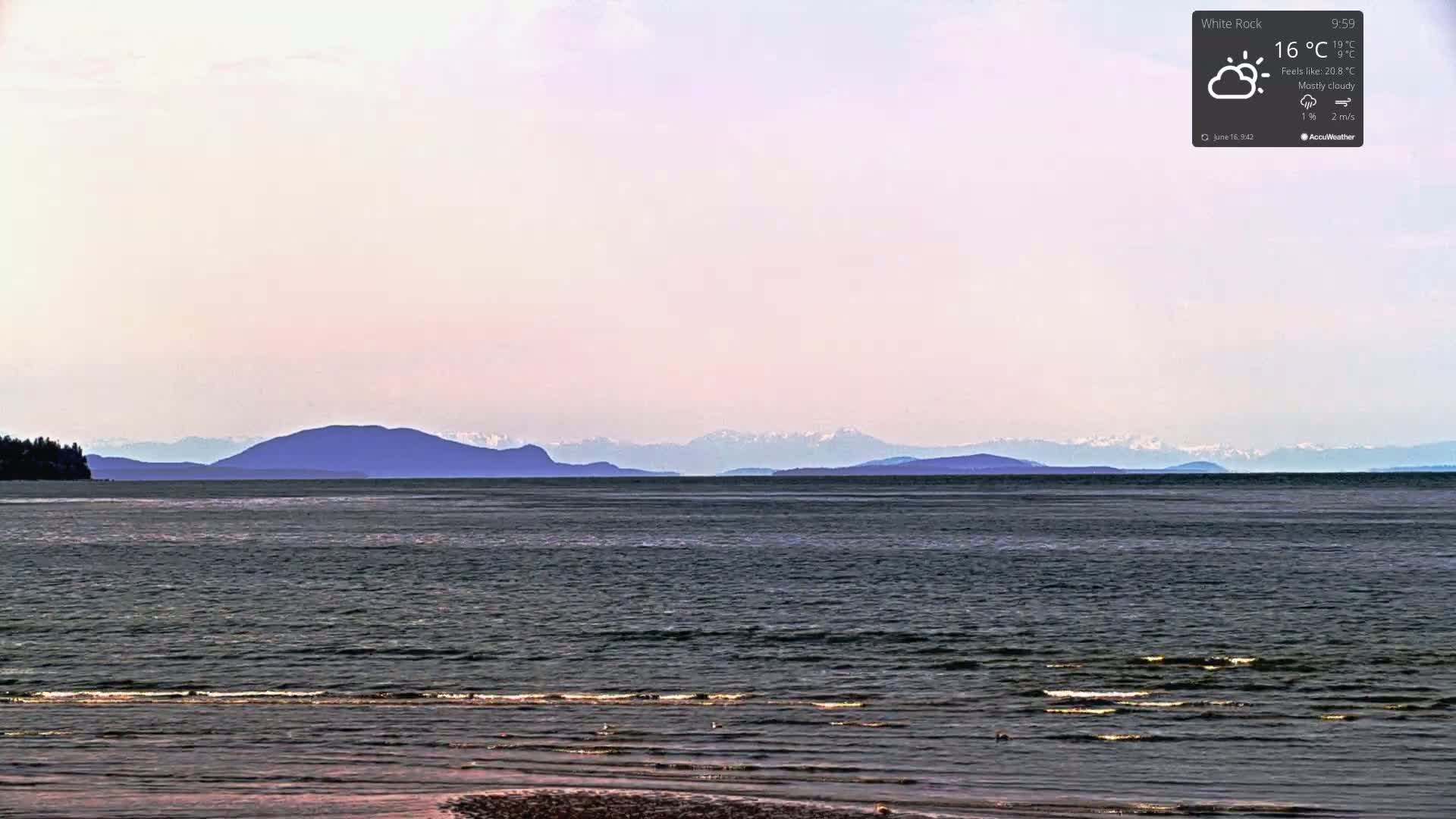 A mostly cloudy, calm day shows a dark body of water leading to distant mountains, some with snow-capped peaks.