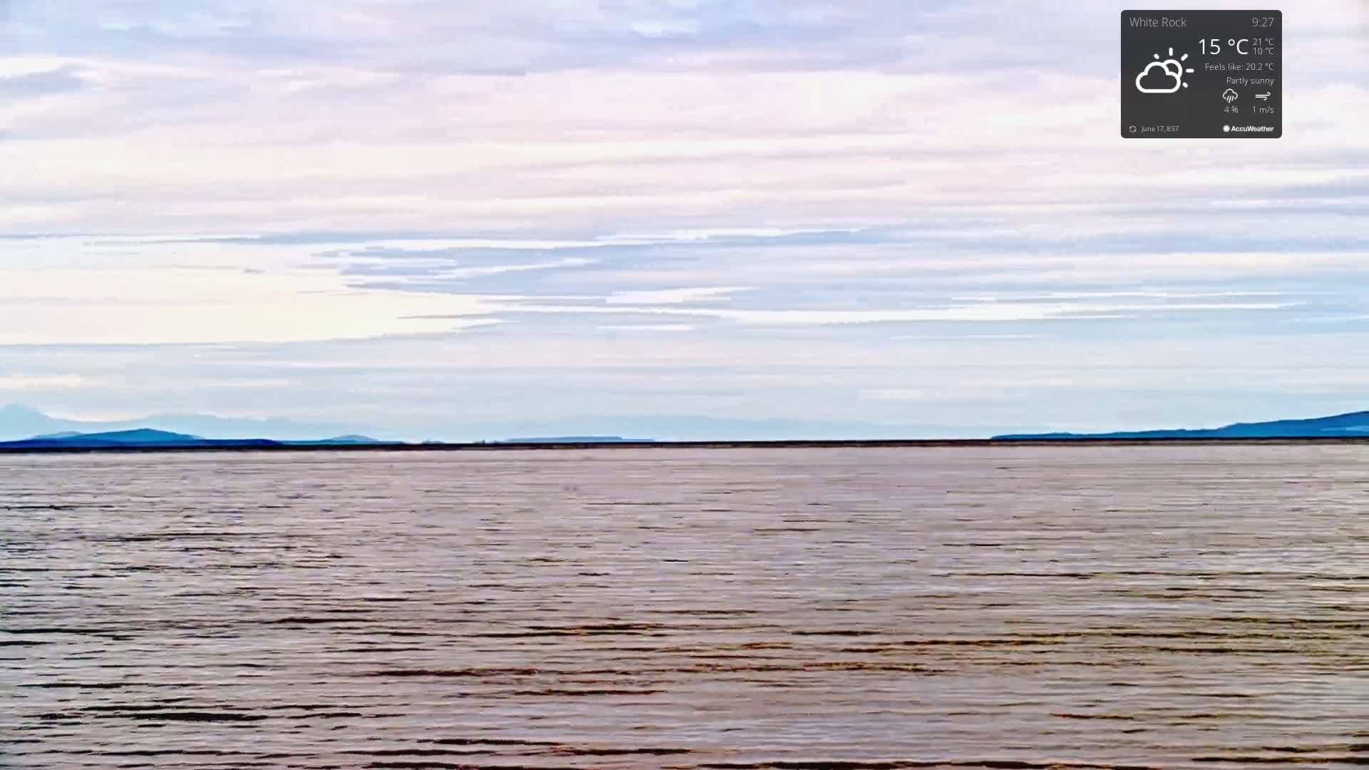 A calm body of water stretches to a distant, low-lying mountain range under a partly sunny sky.