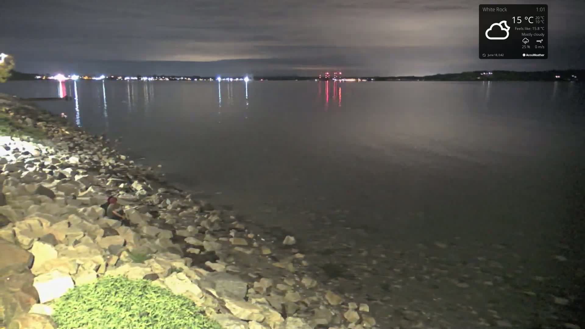At night under mostly cloudy skies, a rocky shoreline curves gently into a calm body of water reflecting distant lights.