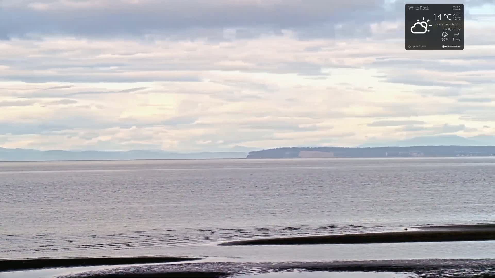 A partly sunny day shows a calm body of water with a distant shoreline and mountains under a mostly cloudy sky.