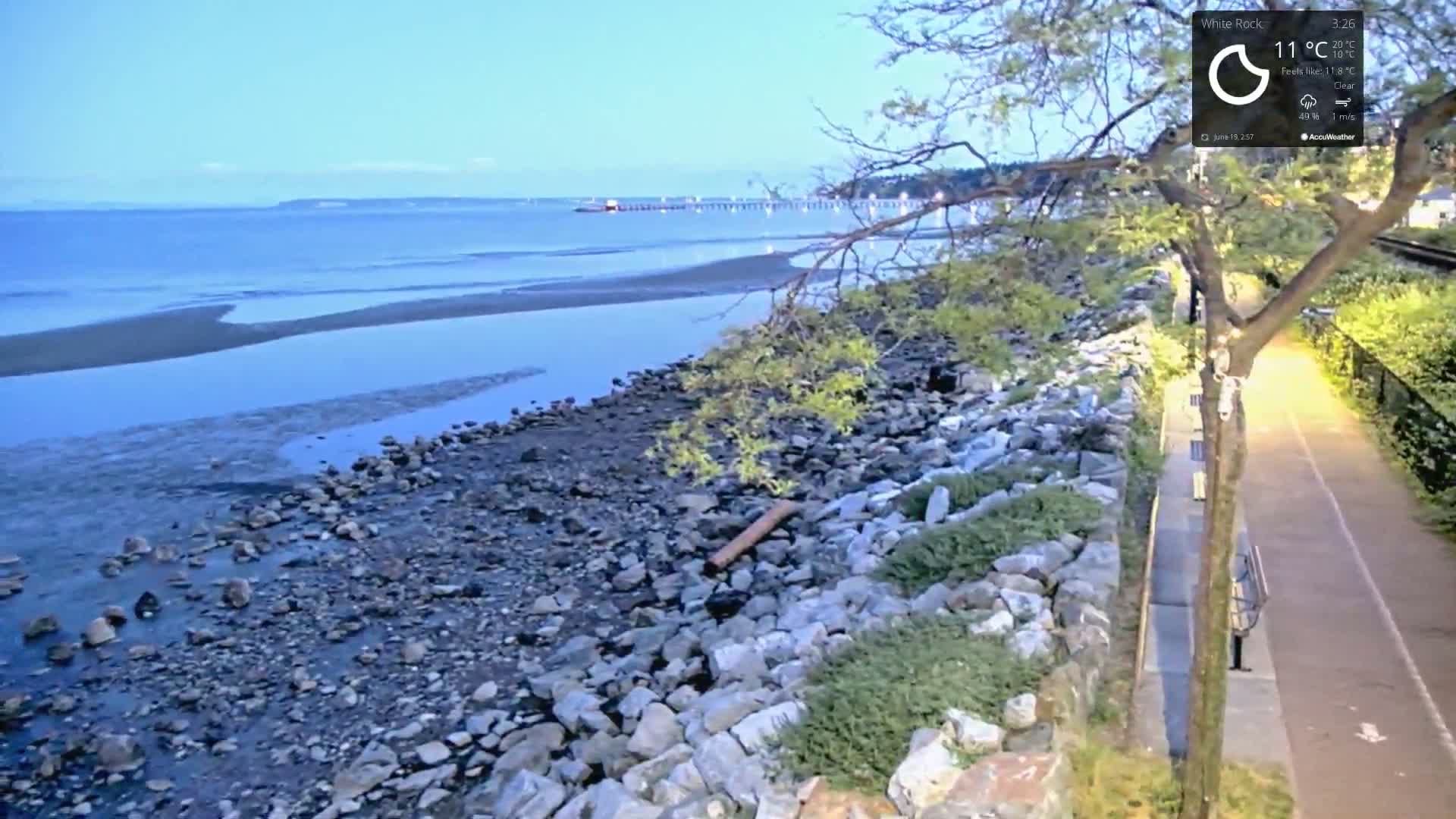 A rocky shoreline with calm water under a clear sky meets a paved path lined with trees and benches.