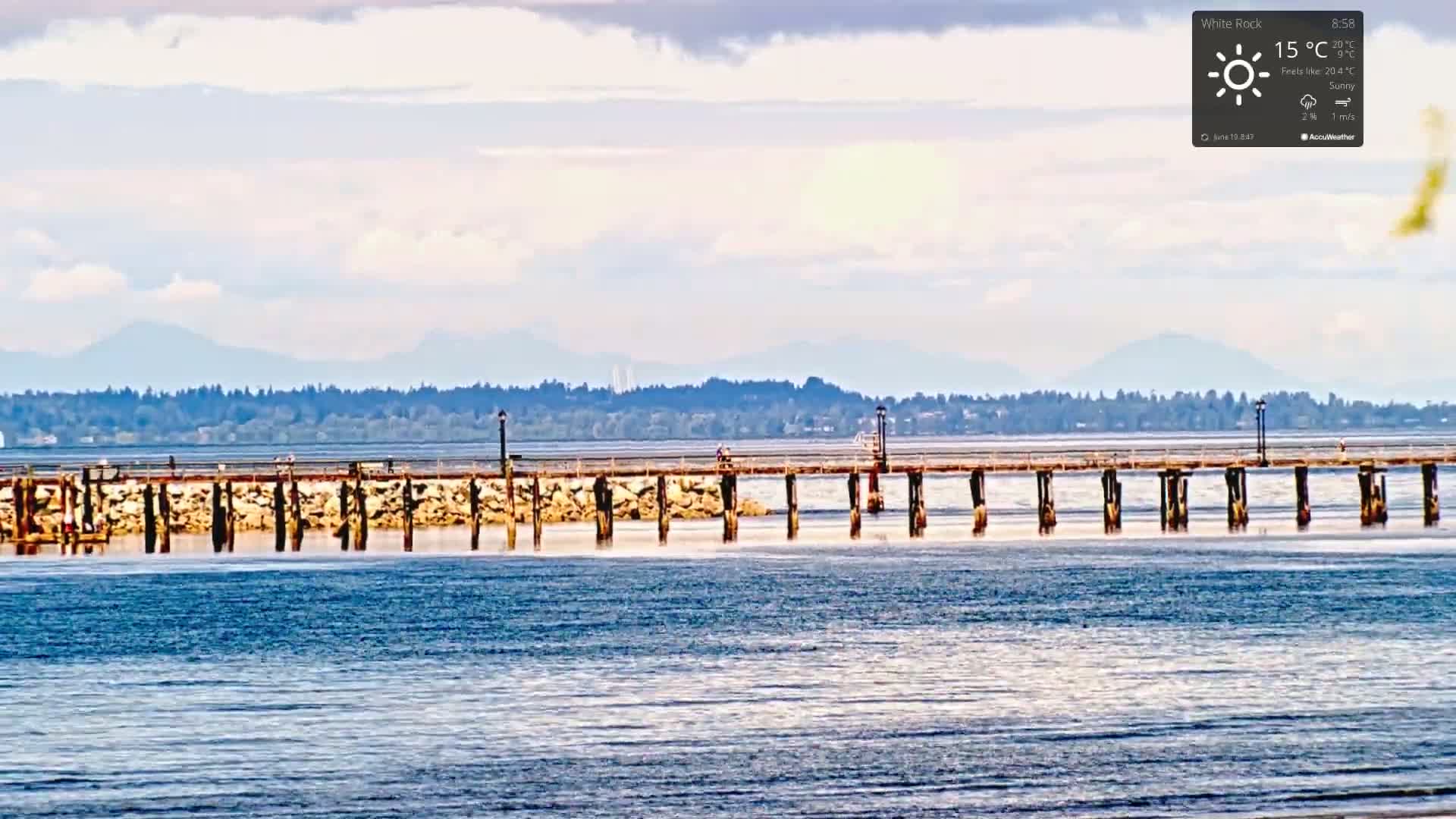 A long pier extends over calm water, with a distant mountain range visible under a partly cloudy sky.