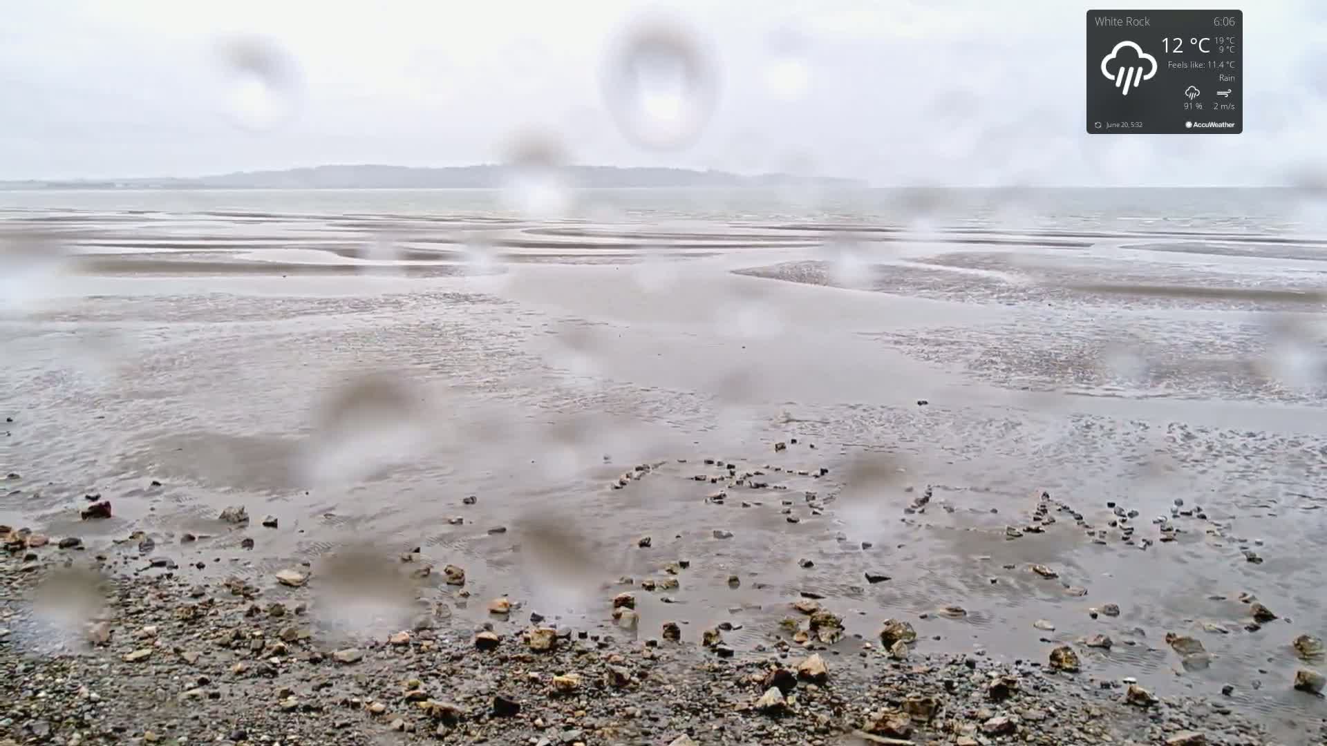 A rain-covered view shows a rocky shore and a tidal flat with rocks arranged to spell out a word on the sand, a distant shoreline visible in the background.