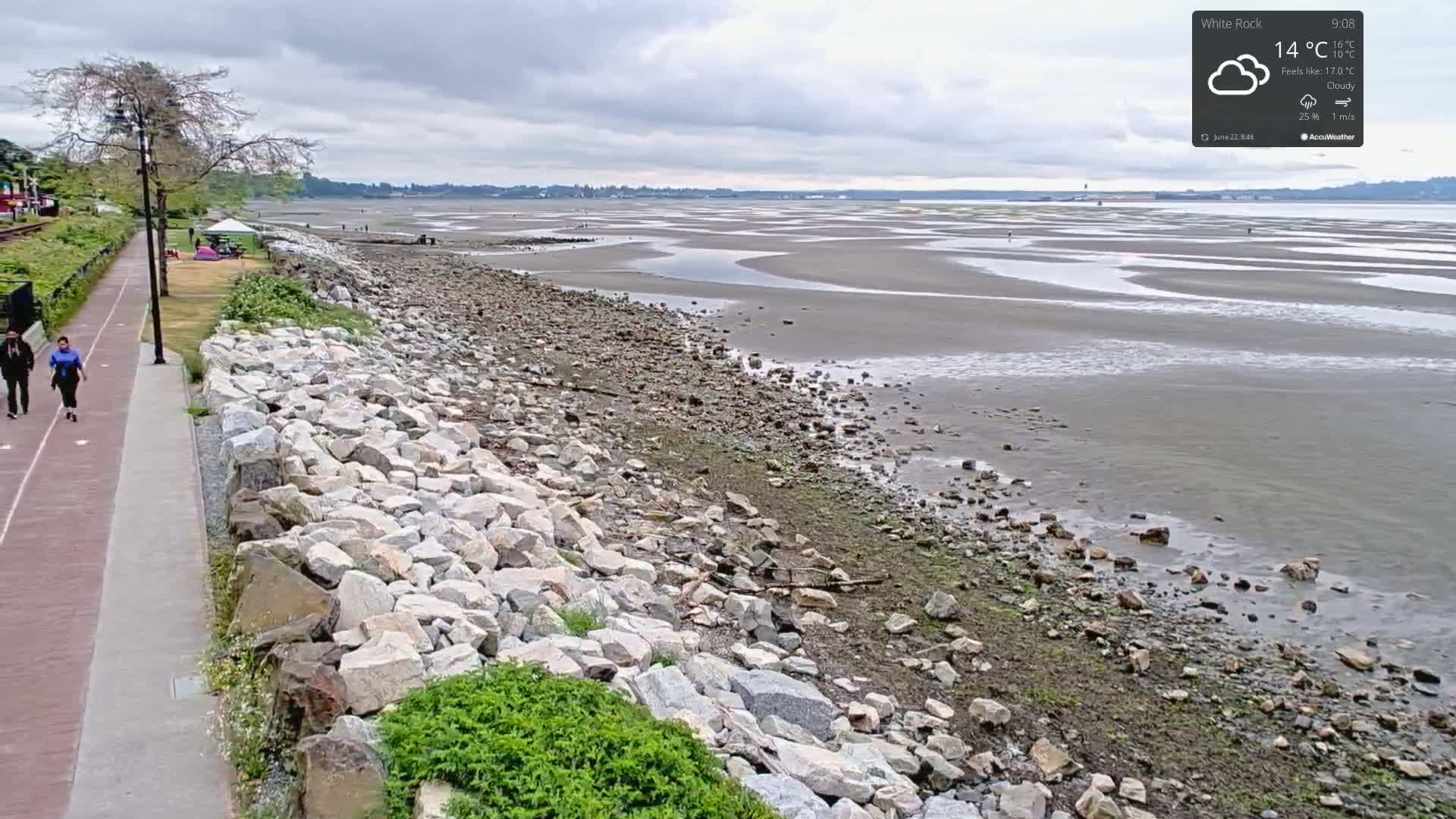 A paved walkway next to a rocky shoreline overlooks a wide expanse of exposed mudflats under a cloudy sky.