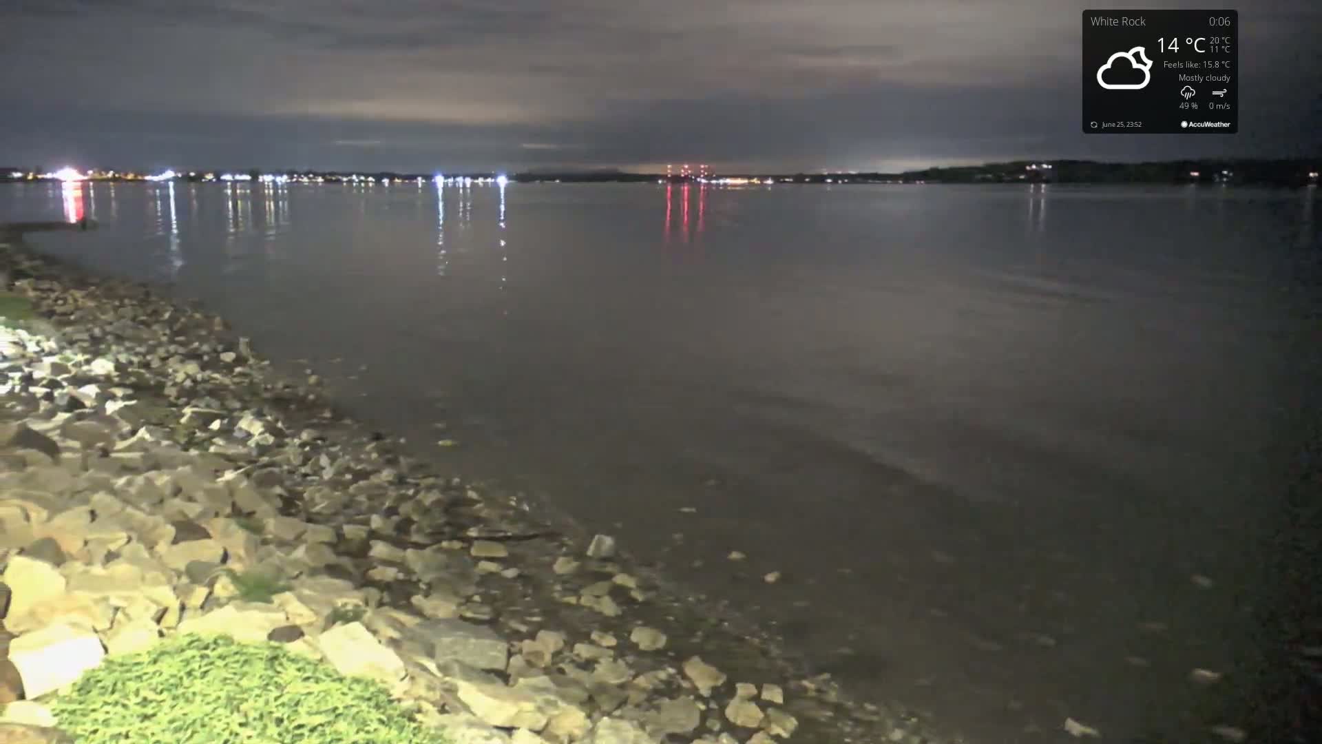 A rocky shoreline at night under mostly cloudy skies overlooks a calm body of water reflecting distant city lights.